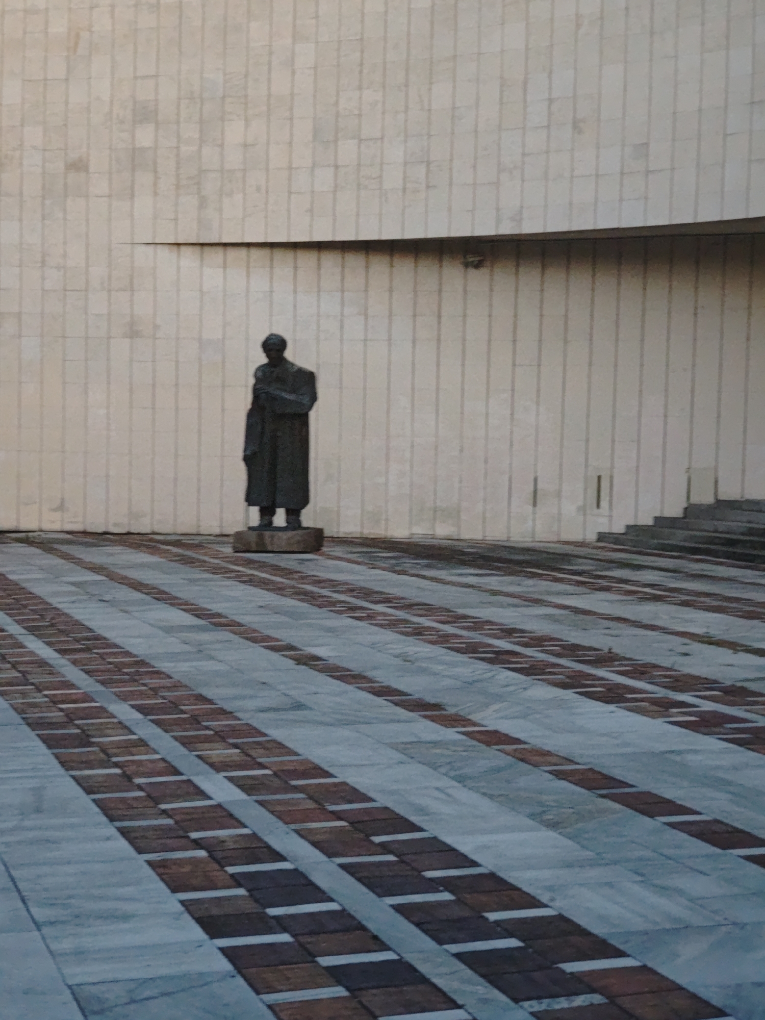 A full-body, standing bronze statue of a man in a long coat and hat is positioned in the center of an open plaza. The plaza's ground is paved with alternating bands of light gray and reddish-brown rectangular tiles, arranged in parallel rows running diagonally away from the viewer. Behind the statue, a textured, light-colored wall extends upwards and to the right, featuring vertical grooves. A dark, angled architectural element cuts across the upper portion of the wall. To the far right of the wall, a set of concrete stairs leads upwards. The lighting suggests it is daytime, possibly late afternoon, casting soft shadows. The statue is located in front of the Regional Historical Museum "Paraskev Tsvetkov" in Pleven, Bulgaria.