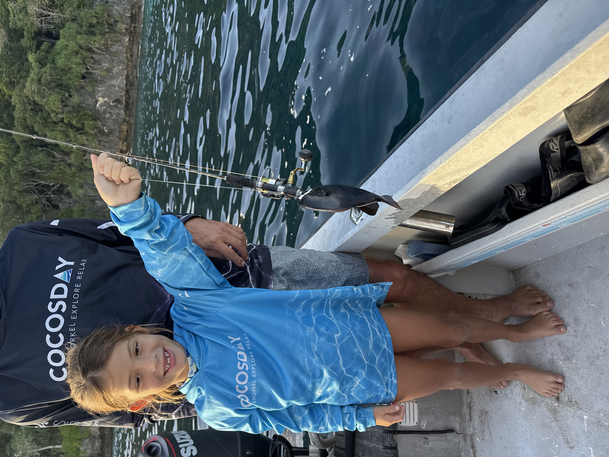 A young girl, her face beaming with joy and a hint of pride, is the central subject of this media file. She is dressed in a long-sleeved blue rash guard with a water ripple pattern and the text "COCOS DAY SNORKEL EXPLORE RELAX" printed on the front. She is holding a fishing rod, with a small, dark fish at the end of the line. Beside her, an adult, likely her father, is partially visible, wearing dark shorts with a white pattern. They are on a boat, evidenced by the white railing and the deck. The scene is set in a body of water, likely the ocean, with lush green trees and a rocky cliff face visible in the background, hinting at a coastal or island location. The water is calm with sunlight reflecting off its surface, suggesting a sunny day, likely in the late afternoon given the warm light. The overall mood is cheerful and adventurous, capturing a moment of family fun and a successful fishing trip. The visible text "SUZUKI" on a boat engine part further confirms the setting is on a boat. The location context of Flying Fish Cove, Australia, further enhances the tropical and adventurous feel of the image.
