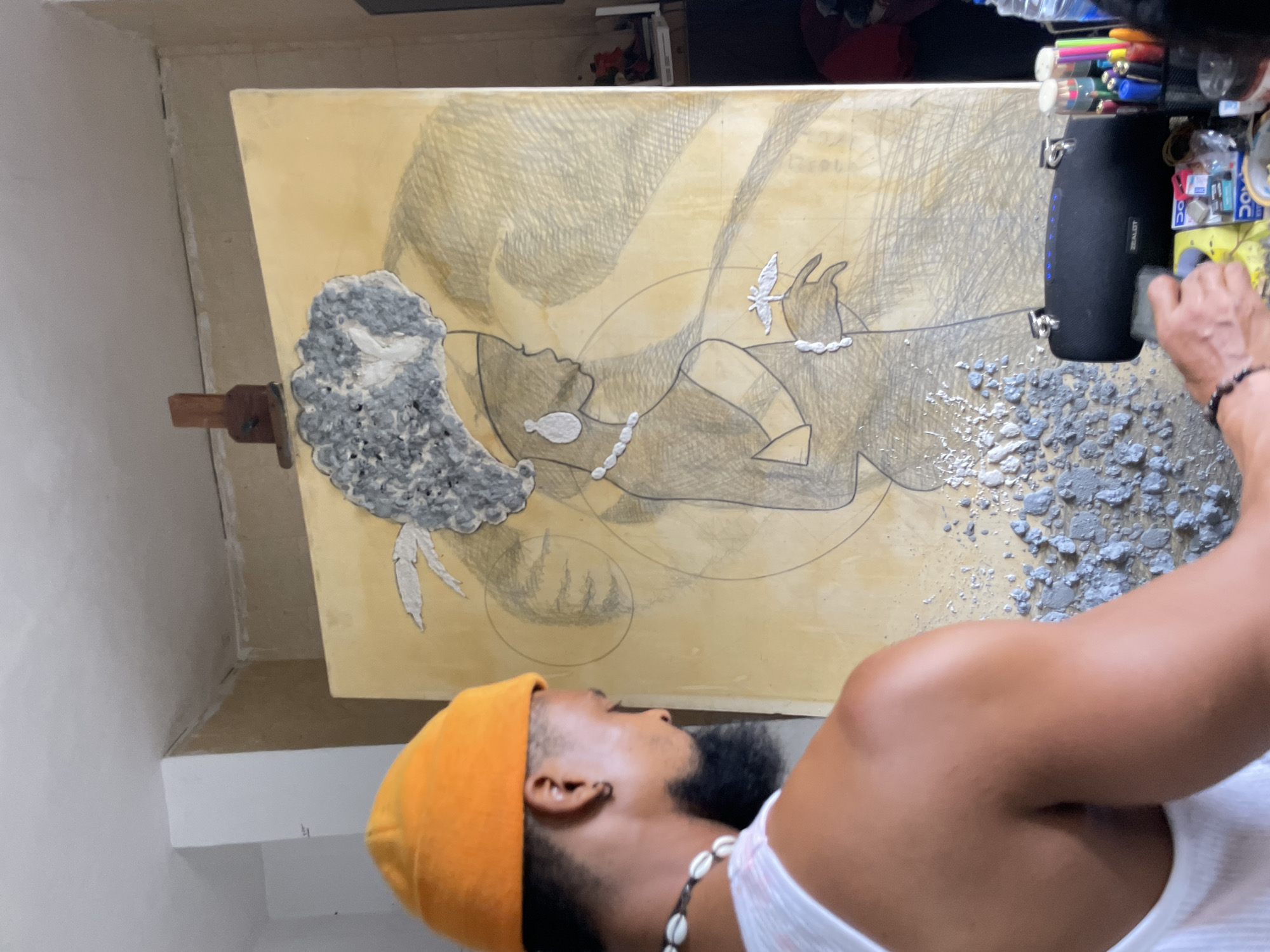 The image shows an artist in Ado, Nigeria working on a piece of art. The artist, a Black man with a beard, wearing an orange beanie and a shell necklace, stands in the foreground. He is close to the canvas, and his arm is visible as he works on it. The artwork is a large canvas on an easel with a yellow background. It features a sketched outline of a figure with a large afro and a necklace. Some areas of the figure, particularly the afro and parts of the clothing, are filled in with a grey, textured material. A black speaker is placed on the canvas, with some art supplies clustered around it, and some of the grey material is scattered nearby. The room appears to be a studio, with white walls and an indistinct background.