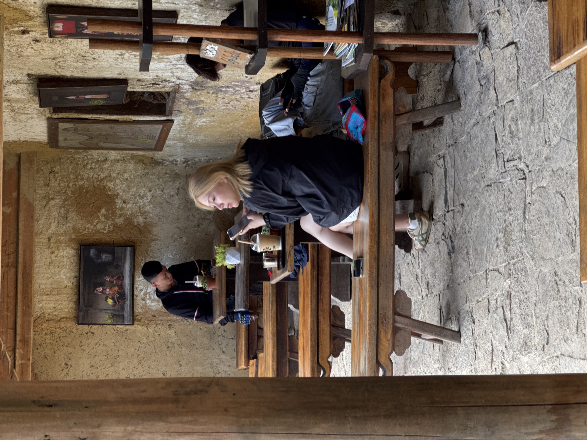 The image depicts an indoor scene in Lũng Cú Commune, Vietnam, with three people seated at adjacent wooden tables. A blonde woman wearing a black jacket and white shorts sits closest to the camera, using a mobile phone. A man in a black jacket is seated next to her, holding a drink. Further down the line of tables is another person, seated and engaged in their own activity. The tables are set with drinks and plants. The walls are a textured tan color, adorned with several framed pictures. The floor is made of grey stone. The shot is taken from a slightly elevated vantage point, partially framed by a wooden structure in the foreground.