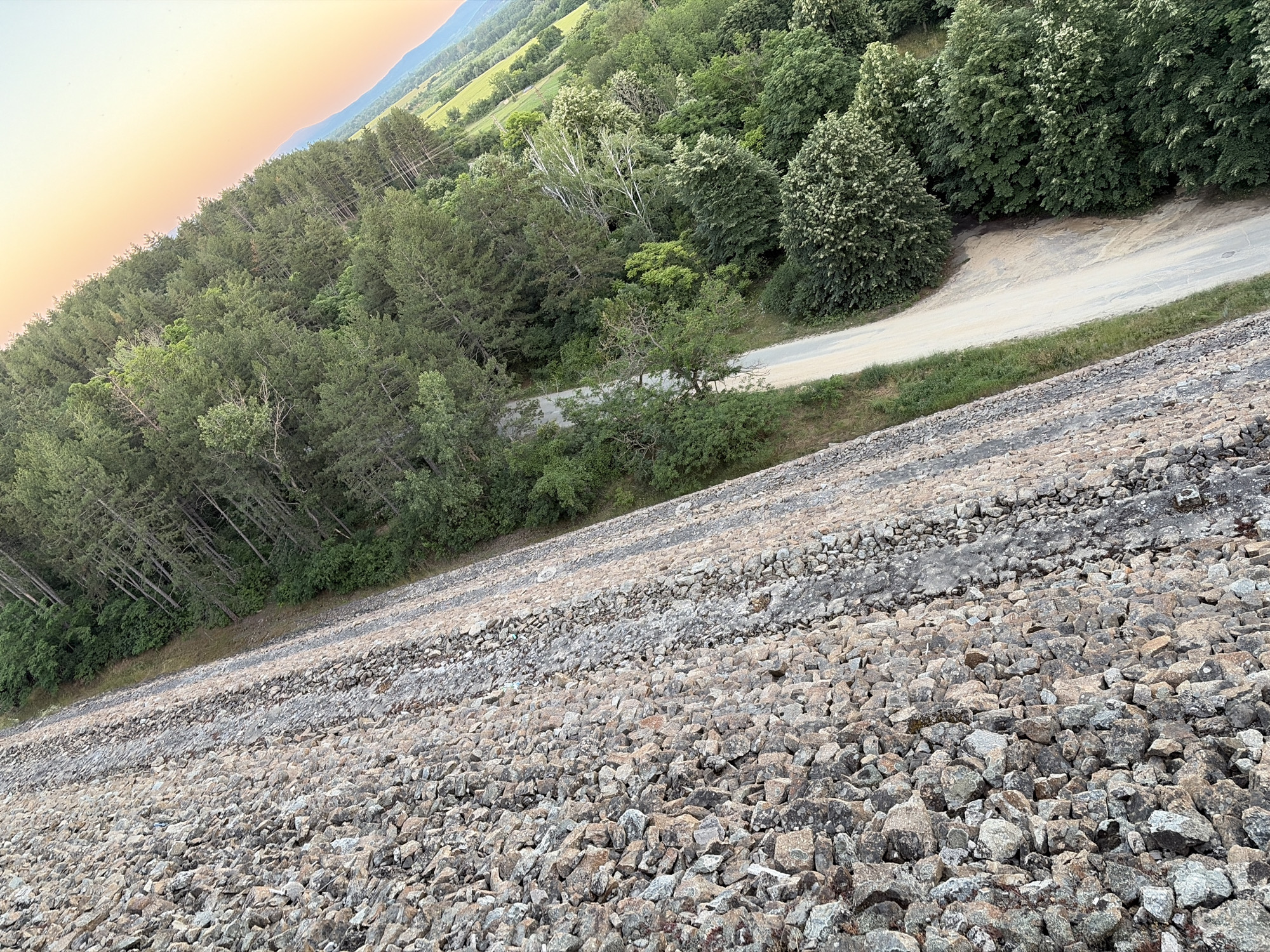 The image depicts a perspective from a high vantage point looking down at a gravel embankment and a forested area with a dirt road winding through it. The embankment is covered in numerous small to medium-sized rocks of varying colors, including gray, brown, and tan. Adjacent to the embankment, a strip of grass and vegetation is visible, followed by a dense forest. The trees are predominantly evergreen, with a mix of lighter and darker green foliage. A dirt road, light in color and appearing somewhat dusty, curves through the trees, disappearing into the distance. The sky above transitions from a pale orange and yellow near the horizon to a light blue, suggesting it may be dawn or dusk. In the far distance, a mountain range can be seen silhouetted against the sky. The image captures the edge of the Koprinka dam in Koprinka, Bulgaria, showcasing its rocky construction and the natural surroundings.