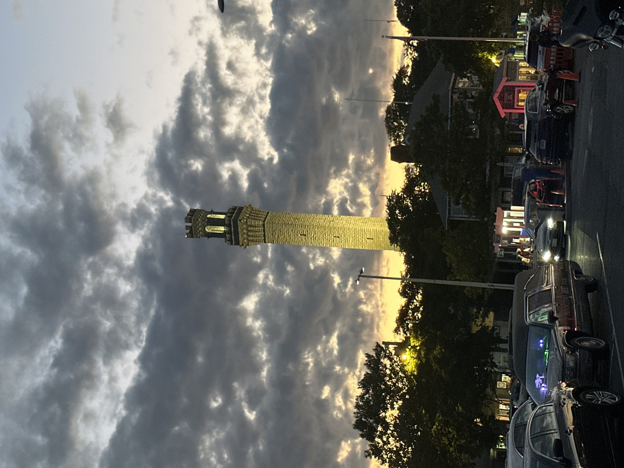 The image shows the Pilgrim Monument in Provincetown, Massachusetts, at dusk. The monument, a tall stone structure, is illuminated and stands prominently against a dramatic sky filled with dark, cloudy formations interspersed with patches of yellow and orange light from the setting sun. In the foreground, a parking lot is visible, with several cars parked. One car, a black hearse, is particularly noticeable, decorated with green and purple lighting inside. Other vehicles, including SUVs and sedans, are also present. Trees and some buildings are visible behind the cars, silhouetted against the twilight sky. A streetlamp is also visible. The overall scene suggests a late evening in a public area, possibly a tourist destination given the monument's presence.