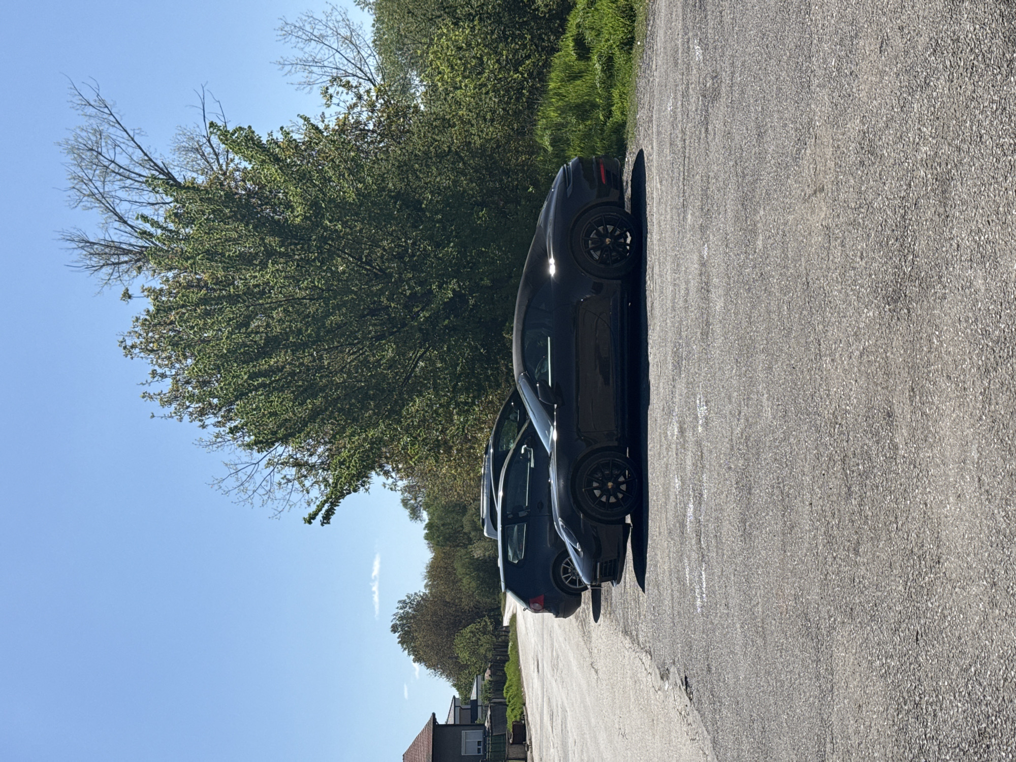The image, oriented correctly, captures a sunlit, tree-lined road with two dark-colored cars parked on its side. The foremost car appears to be a black or dark grey Porsche Macan, identified by its distinct wheel design and partially visible taillight. Another similar dark-colored vehicle is parked directly behind it, partially obscured.
The road surface is made of coarse, grey asphalt, showing some faint white markings. To the left of the parked cars, a dense line of lush green trees, some with full foliage and others with bare branches, extends along the roadside and into the background. The sky above is a clear, bright blue with a few thin, wispy clouds.
Strong sunlight illuminates the scene, casting noticeable shadows from the vehicles onto the road, indicating a daytime setting, likely mid-morning or afternoon with fair and sunny weather. In the distant background on the left, hints of residential buildings with reddish-brown roofs are visible amongst the trees.
The scene depicts a quiet, possibly suburban or semi-rural setting near Sofia, Bulgaria. No specific activity is taking place; the cars are stationary. There is no visible text in the media file.