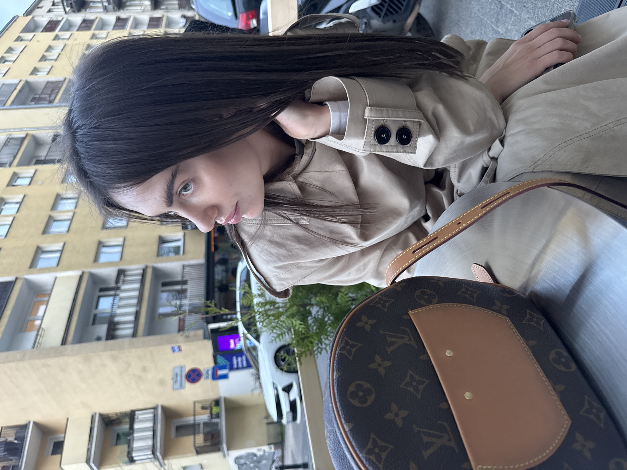 A woman sits outdoors in Warsaw, Poland, wearing a beige trench coat with the sleeves cuffed to reveal a white undershirt. She is next to a Louis Vuitton handbag, with brown monogrammed leather trim visible. Her hair is long and dark, and her gaze is directed towards the left.
In the background, a yellow building with multiple balconies and windows is visible, along with street signs and a white car. The scene is oriented vertically, likely captured from a medium distance.