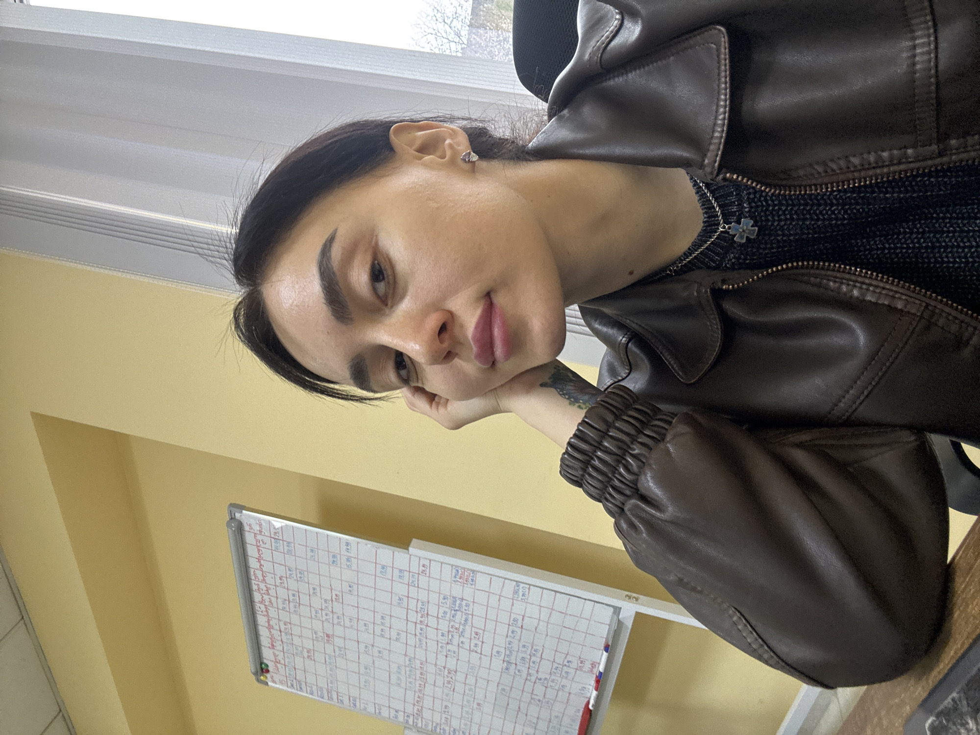 A person is seated indoors, resting their head on their hand. They are wearing a brown leather jacket over a dark knitted sweater, and a necklace with a cross pendant is visible. The person has dark hair pulled back and a small earring in their left ear. A white board with a calendar grid and red writing is visible behind them on a yellow wall. A window with bare trees visible outside is in the background. The location cannot be determined definitively as Tbilisi, Georgia, based on the image.