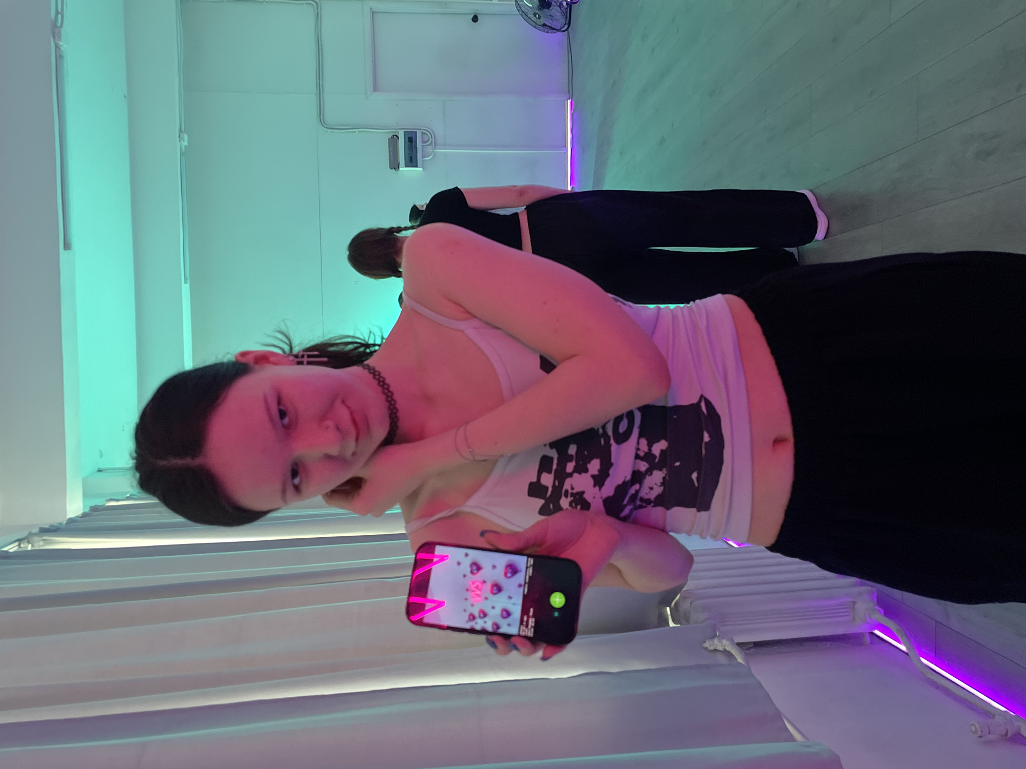 In an indoor setting, likely a studio or practice room in Saint Petersburg, Russia, two young women are illuminated by vibrant artificial lighting that casts green and purple hues across the white walls and light-colored floor. The primary subject, a young woman with dark hair pulled back and wearing a choker necklace, white tank top with a black graphic, and black pants, is in the foreground. She holds a smartphone, tilted slightly, displaying a screen with pink hearts and the text "GM" (Good Morning), alongside a green circular button. She looks towards the viewer with a soft, engaging smile, her arm bent as she holds the phone.
Behind her, another young woman, also dressed in black and with dark hair, stands facing away from the camera. The background reveals white walls with visible electrical conduits, and a floor that appears to be light wood or laminate. On the far left, white blinds or curtains are visible. A fan is partially visible in the upper right corner of the room. The scene has a casual, modern vibe, enhanced by the distinct colored lighting.