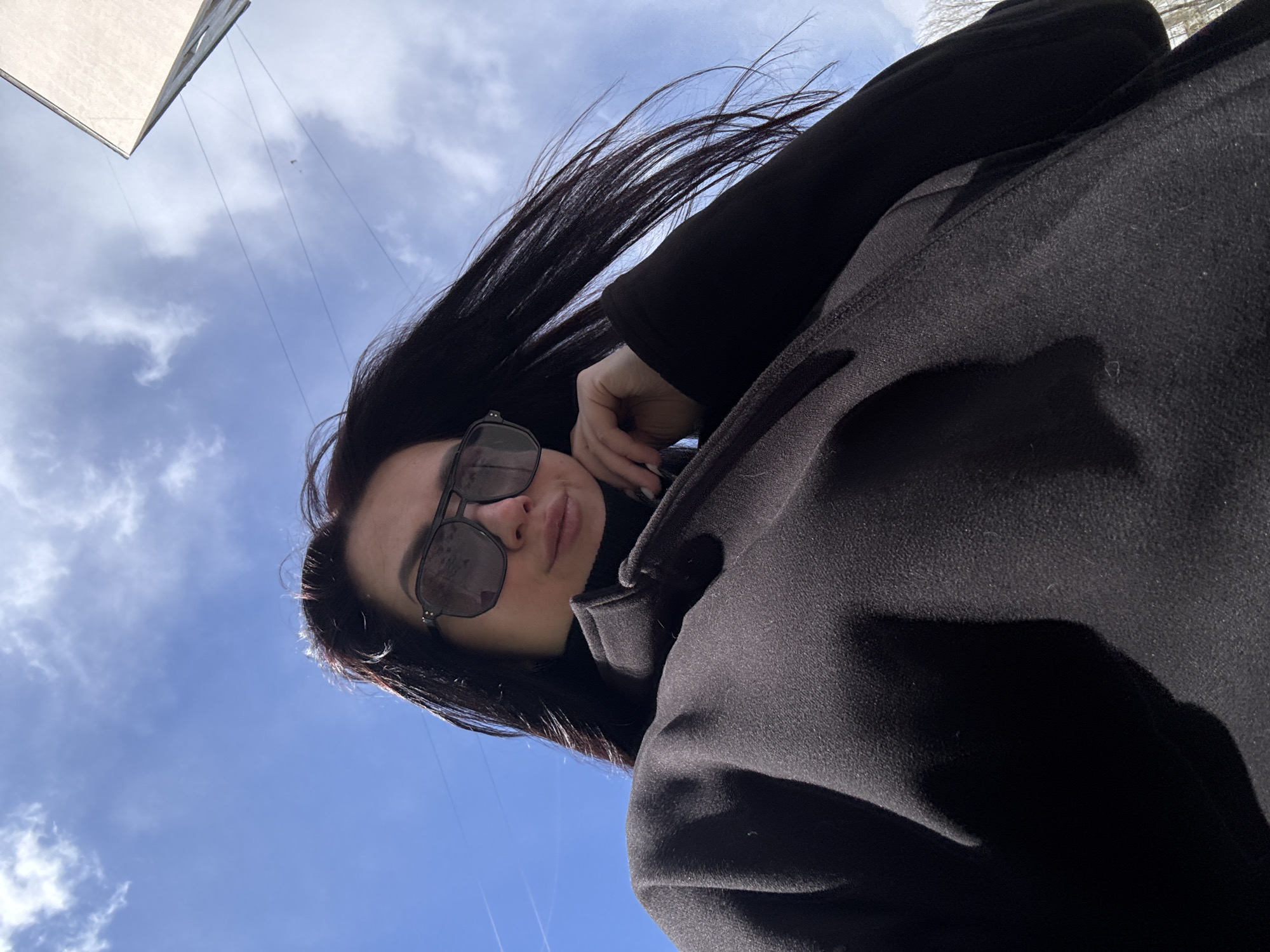 A low-angle, outdoor portrait captures a young woman against a clear blue sky with scattered white clouds. The woman is positioned diagonally in the frame, with her head towards the top left and her body extending towards the bottom right. She wears dark sunglasses and a dark, high-collared garment beneath a black coat or jacket. Her long, dark hair is dramatically swept upwards and to the right, suggesting wind. Her left hand is raised towards her face, resting near her cheek or chin. The bright daylight indicates it is daytime, possibly midday or afternoon, with sunny weather and a noticeable breeze. In the upper left background, a light-colored, angled architectural element, possibly part of a building or roof, is partially visible. Faint lines, which could be wires or aircraft contrails, are also discernible against the sky. Her expression is neutral, largely concealed by the sunglasses. There is no visible text in the image.
