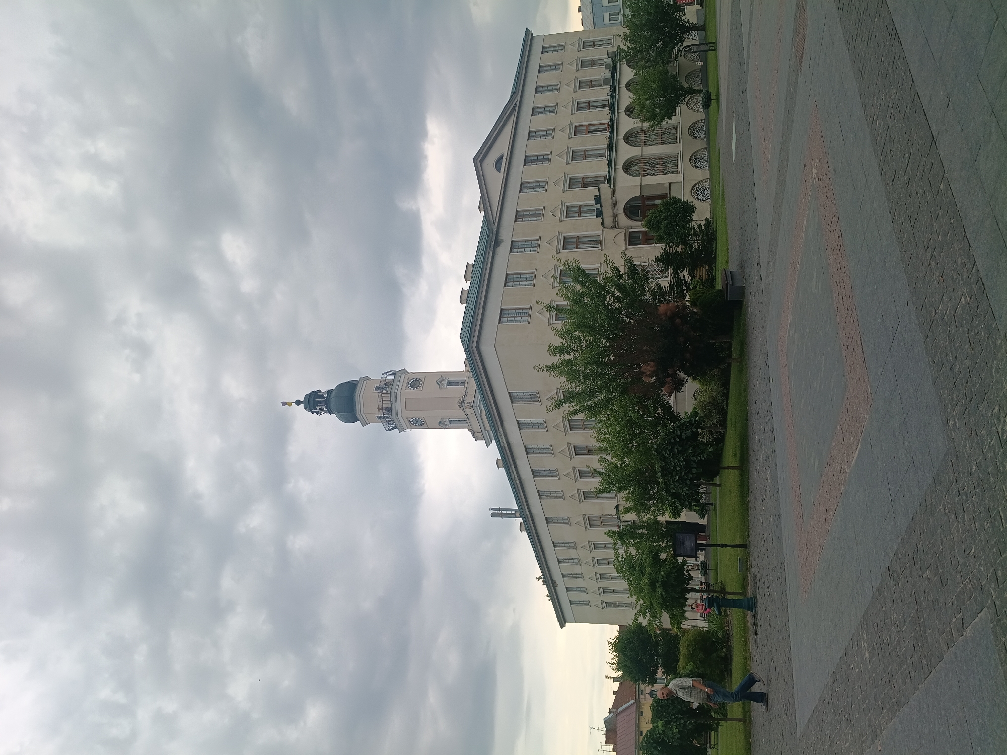 The image shows a cloudy day in Drohobych, Ukraine, featuring a large, light-colored building with a prominent clock tower. The building appears to be a public structure, possibly a town hall or administrative building, with many windows and classical architectural elements. In the foreground, a paved square with intricate stone patterns is visible. A lone man in a plaid shirt and jeans walks across the square, facing left. Several green trees are planted in front of the building, and a fire hydrant is present on the left side of the square. The sky is heavily overcast with grey clouds, suggesting a temperate or possibly humid climate. The overall scene is one of a quiet, urban environment under a somber sky.