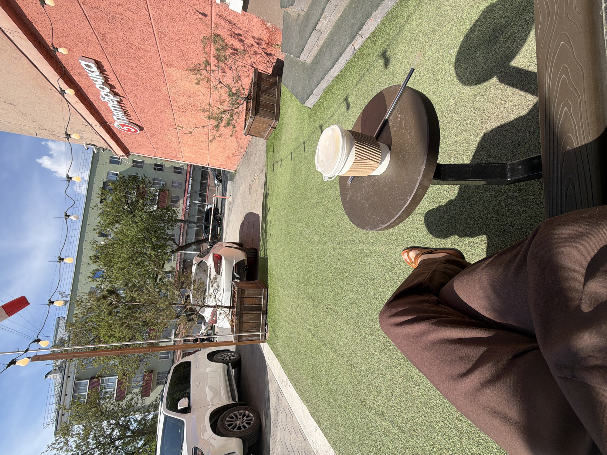The image depicts an outdoor scene in Perm, Russia, featuring a person seated at a table. A disposable coffee cup with a straw sits on the table. The person is wearing brown pants and sandals. The ground is covered in artificial turf. In the background, buildings and parked cars are visible, including the "Pyaterochka" grocery store. String lights hang above the street.