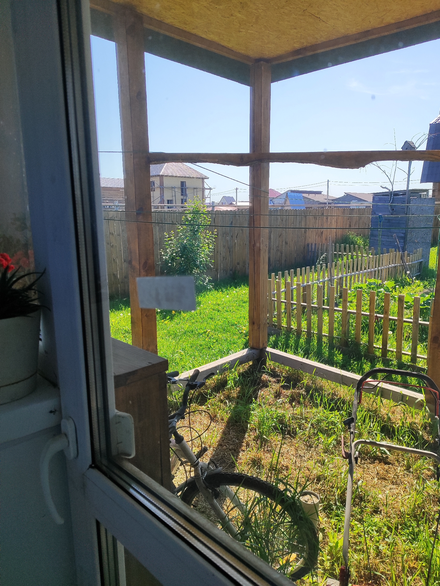 The image shows a partially constructed wooden veranda overlooking a grassy yard. In the foreground, a white window frame with a handle is visible. Inside the veranda, a bicycle with a dark frame is leaning against a wooden structure. A lawnmower with red accents is also present. The yard is enclosed by a wooden fence. Beyond the fence, residential buildings and a clear blue sky are visible. The location is identified as Marusino, Russia.
