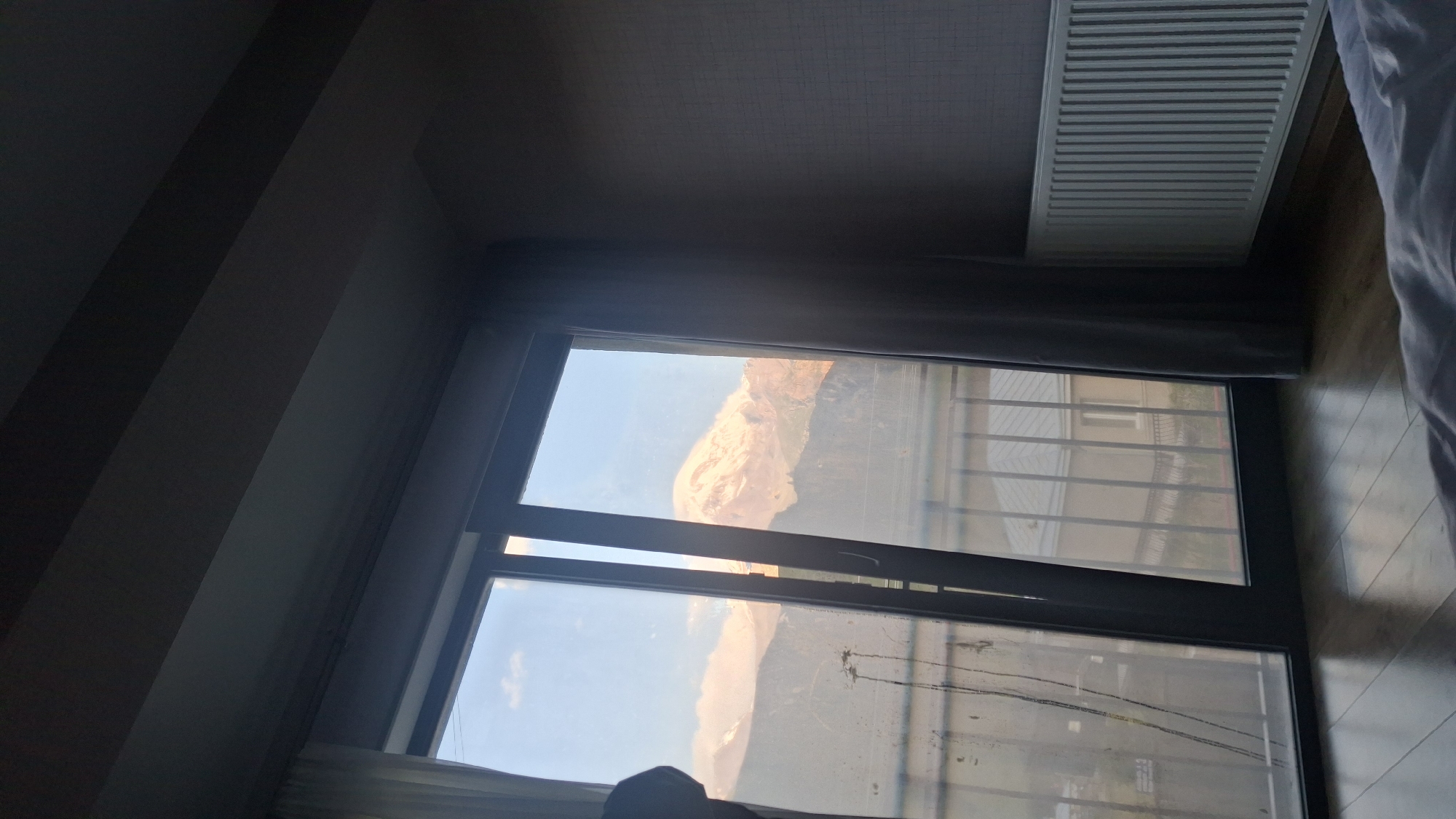 The image shows a view from a room in Stepantsminda, Georgia, overlooking Mount Kazbek. The foreground features a wooden floor and a portion of a bed with white bedding. To the left, a large window with dark framing is visible. The window is divided into two panels, with the left panel slightly ajar. Through the window, a balcony railing is present, and beyond that, a building with a grey roof is seen. The main focus of the view outside is the majestic Mount Kazbek, its snow-capped peak illuminated by the soft light of either sunrise or sunset. The sky is a clear blue with a few wispy clouds around the mountain. The interior of the room is dimly lit, with dark curtains framing the window and a white radiator mounted on the wall to the right. The overall impression is one of a peaceful morning or evening with a stunning natural backdrop.