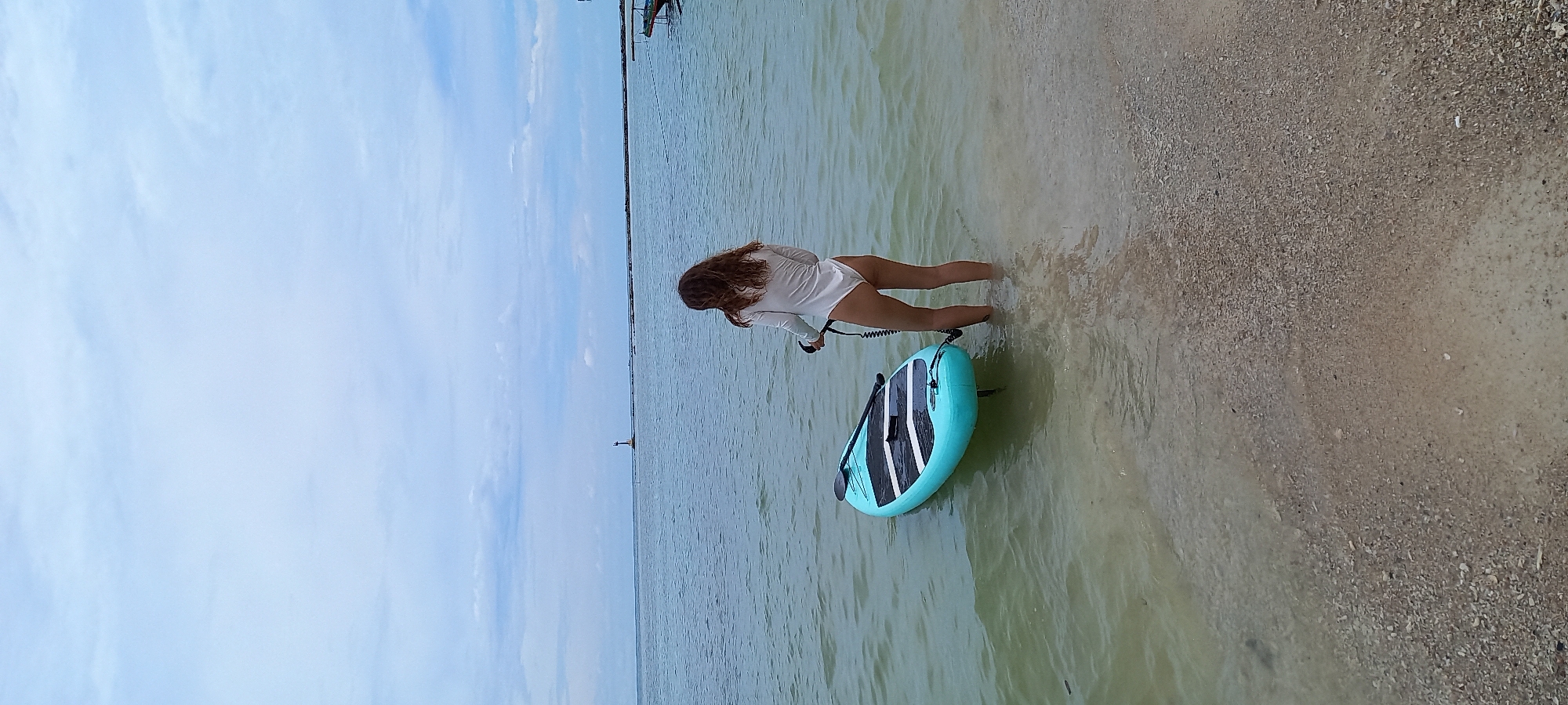 A young woman, waist-deep in shallow, slightly murky water, prepares to use a turquoise and black paddleboard. She is wearing a white long-sleeve shirt. The location appears to be a beach near Baan Chaloklum, Thailand, as evidenced by a distant navigational marker (likely a lighthouse) on the horizon. A strip of land is also visible in the distance. The sky is overcast with streaks of light blue. The beach is composed of light-colored sand and small pebbles.