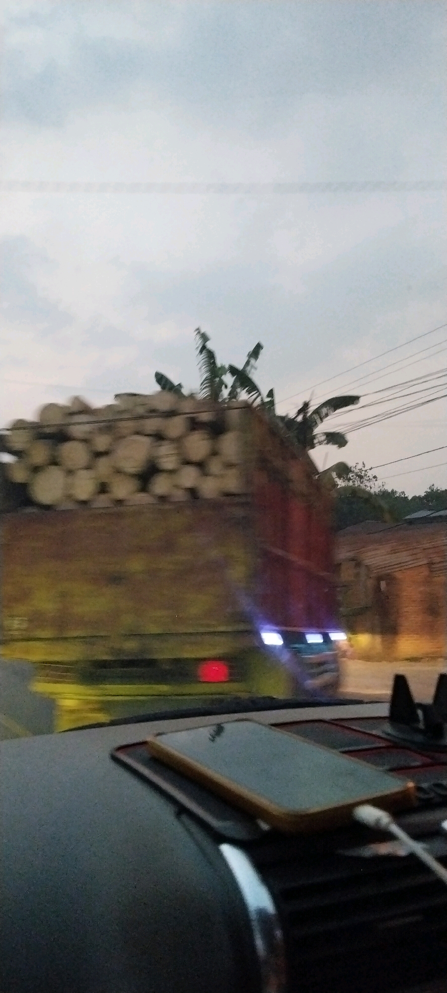 The image captures a motion-blurred scene from inside a vehicle, looking out at a truck loaded with logs. The truck appears to be moving, as indicated by the blur. In the background, there are buildings and trees, with power lines visible against the overcast sky. A smartphone is placed on the dashboard of the vehicle in the foreground. The scene appears to be in Kali Balangan, Indonesia, given the context provided.