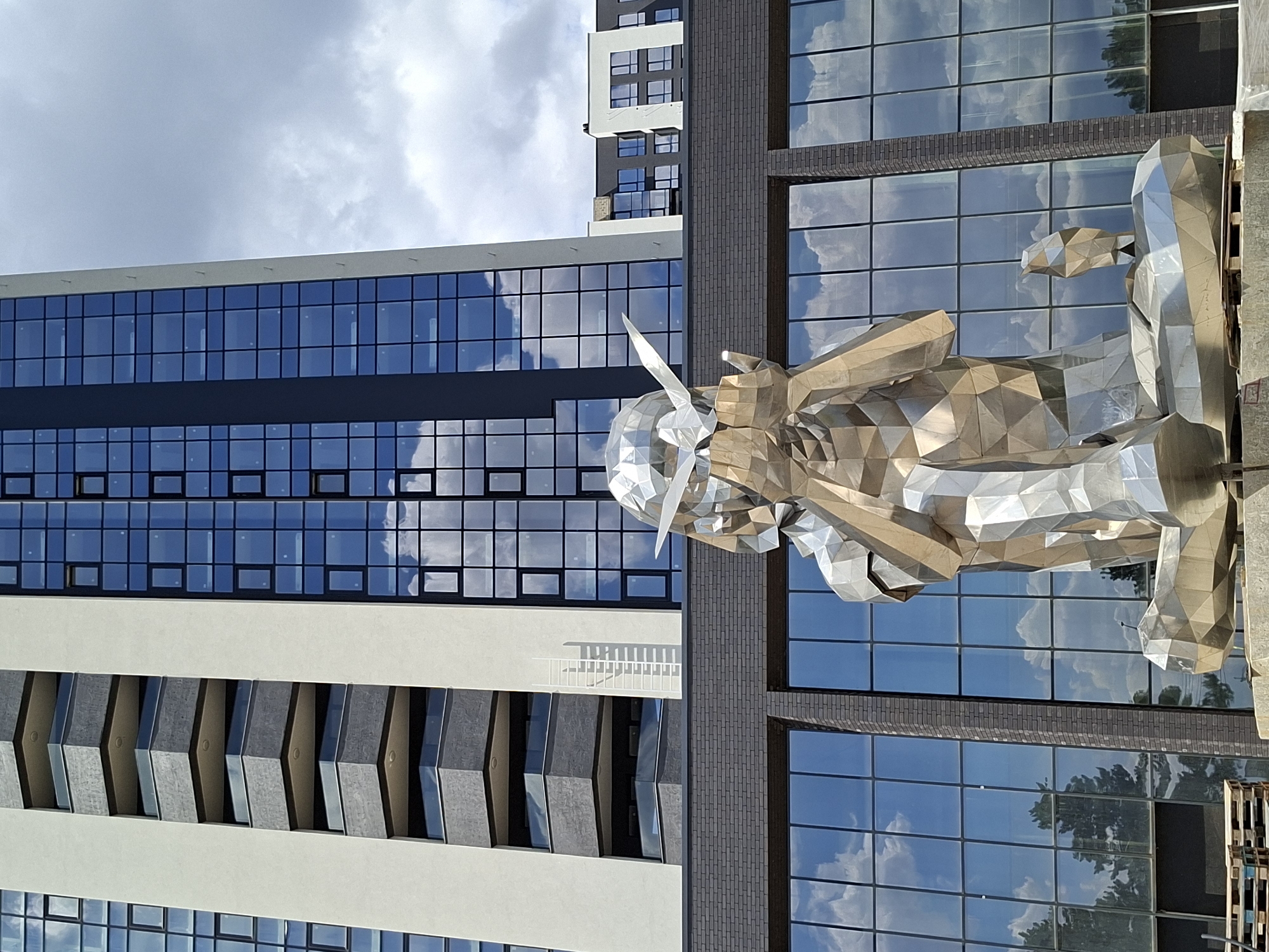 The image shows a modern building with a glass facade and a large, geometric metal sculpture in the foreground. The sculpture depicts a seated figure with its hands covering its face, holding a small object in its right hand. A smaller, abstract sculpture sits to the right of the main figure. The building behind the sculpture features multiple stories with balconies on the left side and a grid of windows on the right. The facade of the lower part of the building is made of dark bricks. The sky above is partly cloudy. The sculpture is located in front of the residential complex "Imagine Residence" in Chișinău, Moldova.