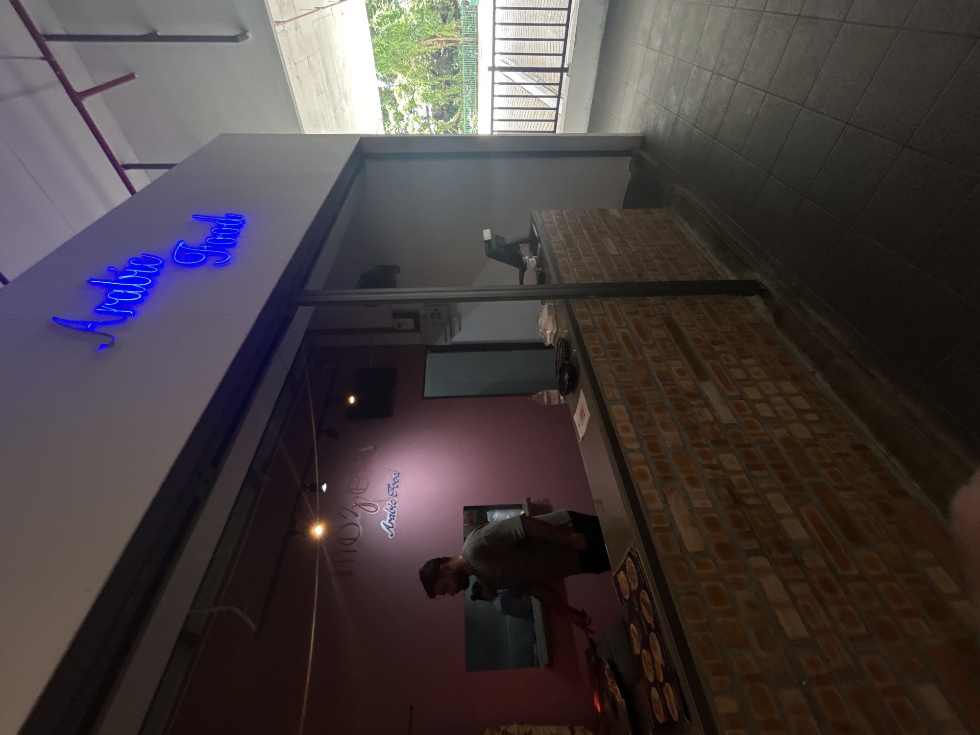 The image captures the interior of a food establishment named "Arabic Food," possibly located in Kuala Lumpur, Malaysia. The scene presents a service counter with a male employee standing behind it, apparently attending to customers. A neon sign advertising the establishment is visible. The counter area is constructed of brick and features menus or displays showing food items. The lighting is a mix of ambient and illuminated, creating contrasts within the space. Outside the establishment, there's a glimpse of a covered walkway and some greenery, suggesting an outdoor area or a connection to a garden or park. The scene presents a casual dining environment.