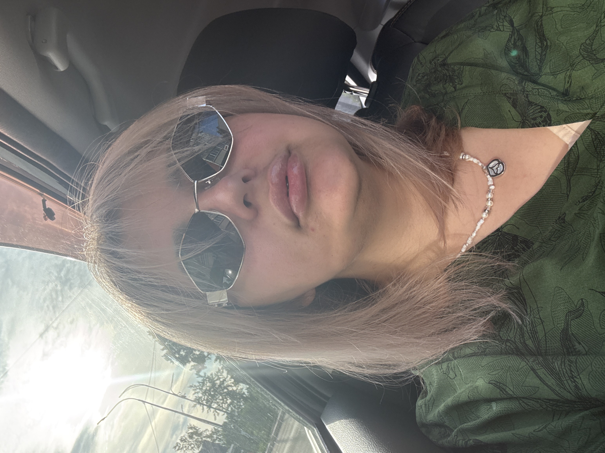 A young woman with blonde hair and sunglasses is in the front passenger seat of a car, possibly in Birobidzhan, Russia, given the provided context. She is wearing a green shirt with a dark floral pattern and a pearl necklace with a silver pendant. The sun is shining brightly through the car's sunroof, casting a glare on her face and obscuring some of the background details. The interior of the car is visible, including the headrest and part of the seat. Outside the car, trees and streetlights can be seen, suggesting an urban or suburban environment.