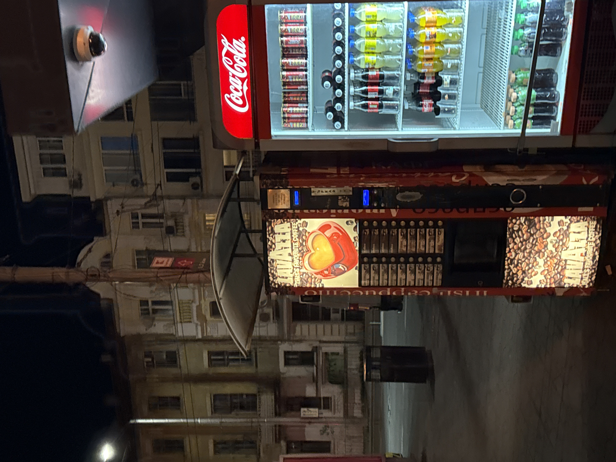 The image depicts a Coca-Cola branded vending machine in Varna, Bulgaria, located outdoors on a city street at night. The machine is positioned in front of a multi-story building with numerous windows. The vending machine is stocked with various beverages, including Coca-Cola, Fanta, and other soft drinks. Below the beverage section is a coffee vending machine, advertising cappuccino and decorated with coffee beans. A surveillance camera is mounted on a nearby structure. A street lamp illuminates the scene, and a trash receptacle is visible nearby. The street appears to be sparsely populated.