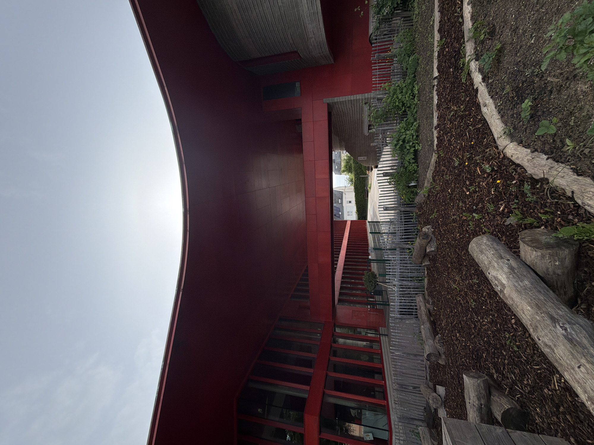 The image depicts a modern architectural structure with a striking red facade, located in Esch-sur-Alzette, Luxembourg. The building has a curved, cantilevered section that creates a covered space. A series of windows and a staircase are visible, suggesting multiple levels within the building. Adjacent to the building is a garden area with wood chip mulch, logs, and small plants. In the background, a glimpse of a street and buildings is visible. The sky is partially overcast.