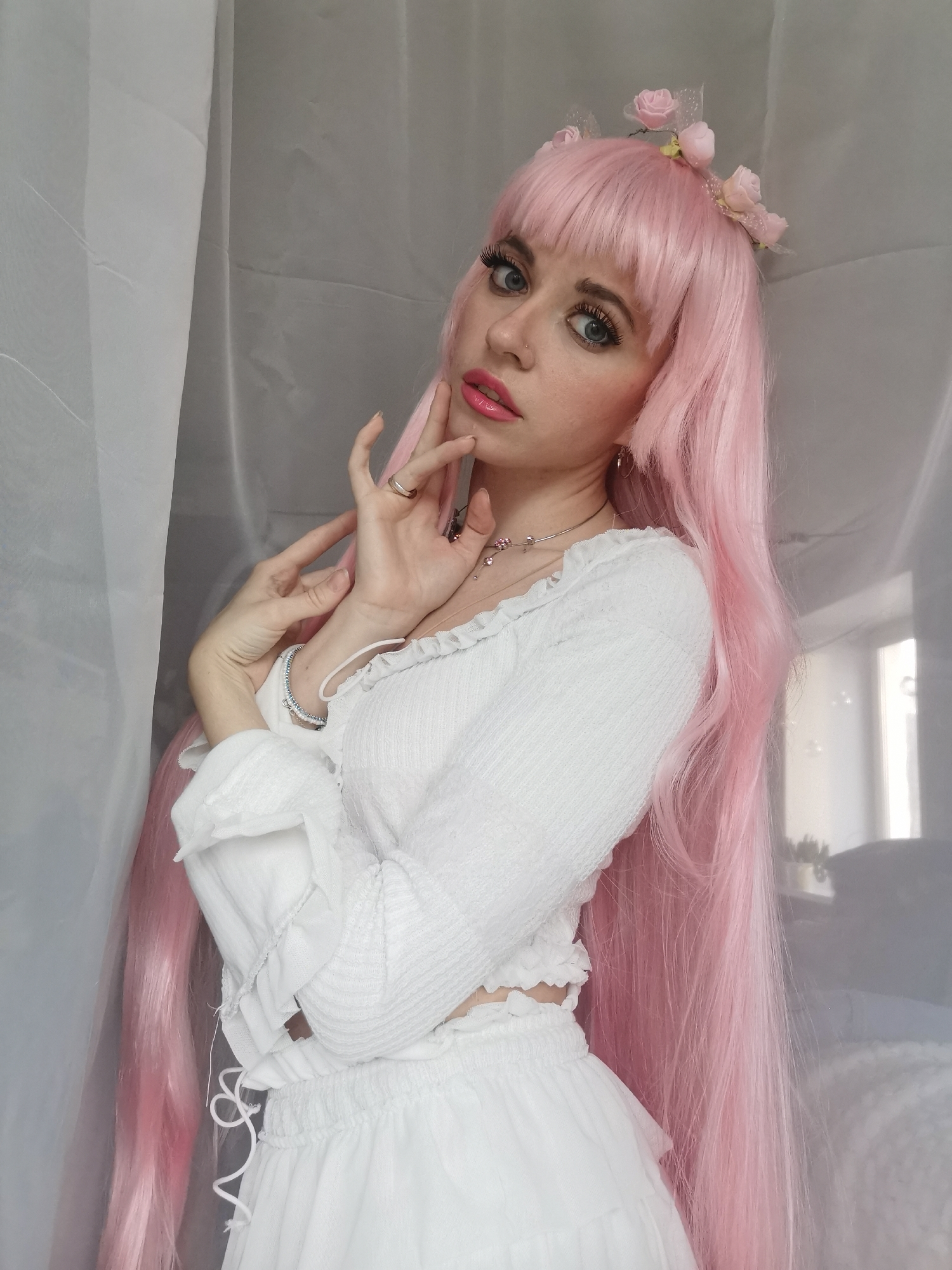 The image captures a young woman posing indoors, looking directly at the viewer with a soft, engaging expression. She has long, vibrant pink hair with bangs, styled with a delicate headband adorned with small pink roses and white netting. Her blue eyes are accentuated with dark eyeliner and long lashes, and her lips are painted with a glossy pink lipstick.
She is dressed in a white, long-sleeved top featuring a textured, possibly smocked, fabric with ruffled details at the neckline and cuffs. A slender, silver-toned necklace with small charms hangs around her neck, and she wears a silver-toned ring on her right ring finger, along with a light-colored bracelet on her left wrist. Her hands are positioned gracefully near her face, with her right hand gently touching her chin. The bottom part of her outfit is a matching white skirt or shorts with a ruffled, smocked waistband and visible drawstrings on the left side.
The background consists of soft, light-colored, possibly sheer, fabric or curtains draped to the left and behind her, creating an ethereal ambiance. To the far right, reflections suggest a window or a significant light source, and some indistinct light-colored furniture, possibly a bed, is faintly visible. The lighting is bright and diffused, indicating daytime. No text is visible in the image.