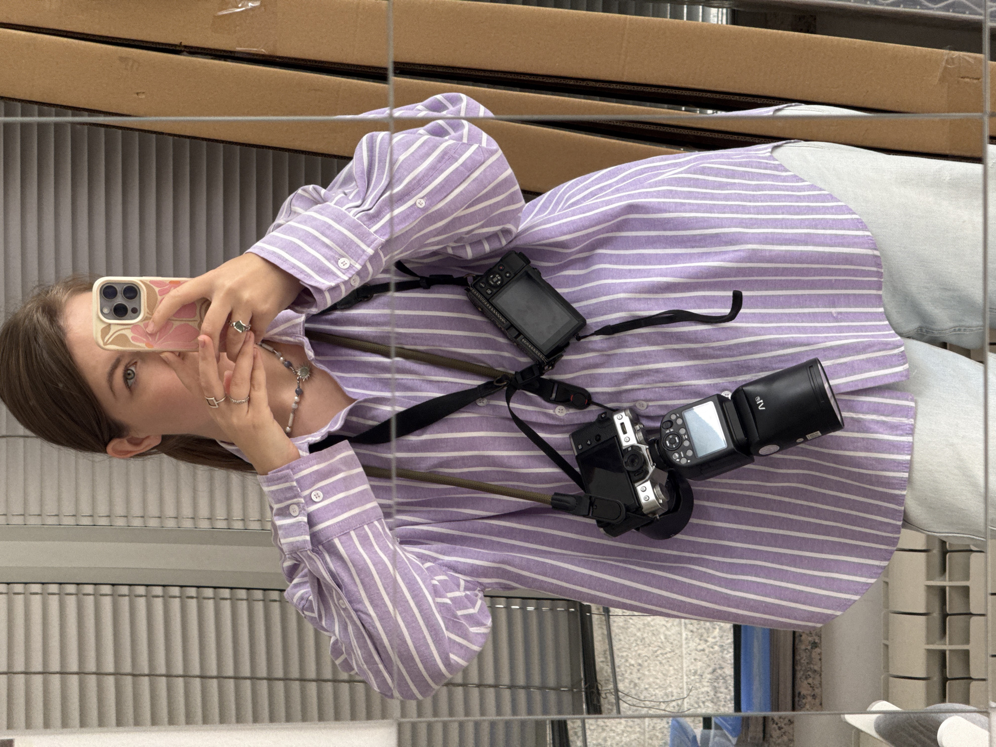 A woman with brown hair stands in front of a mirror, taking a picture of herself with a pink floral-patterned smartphone. She wears a lavender and white striped button-down shirt and light wash denim jeans. A camera with a flash attachment is slung across her chest. The reflection shows the environment includes vertical blinds and cardboard boxes. The location is Vracar Urban Municipality, Serbia.