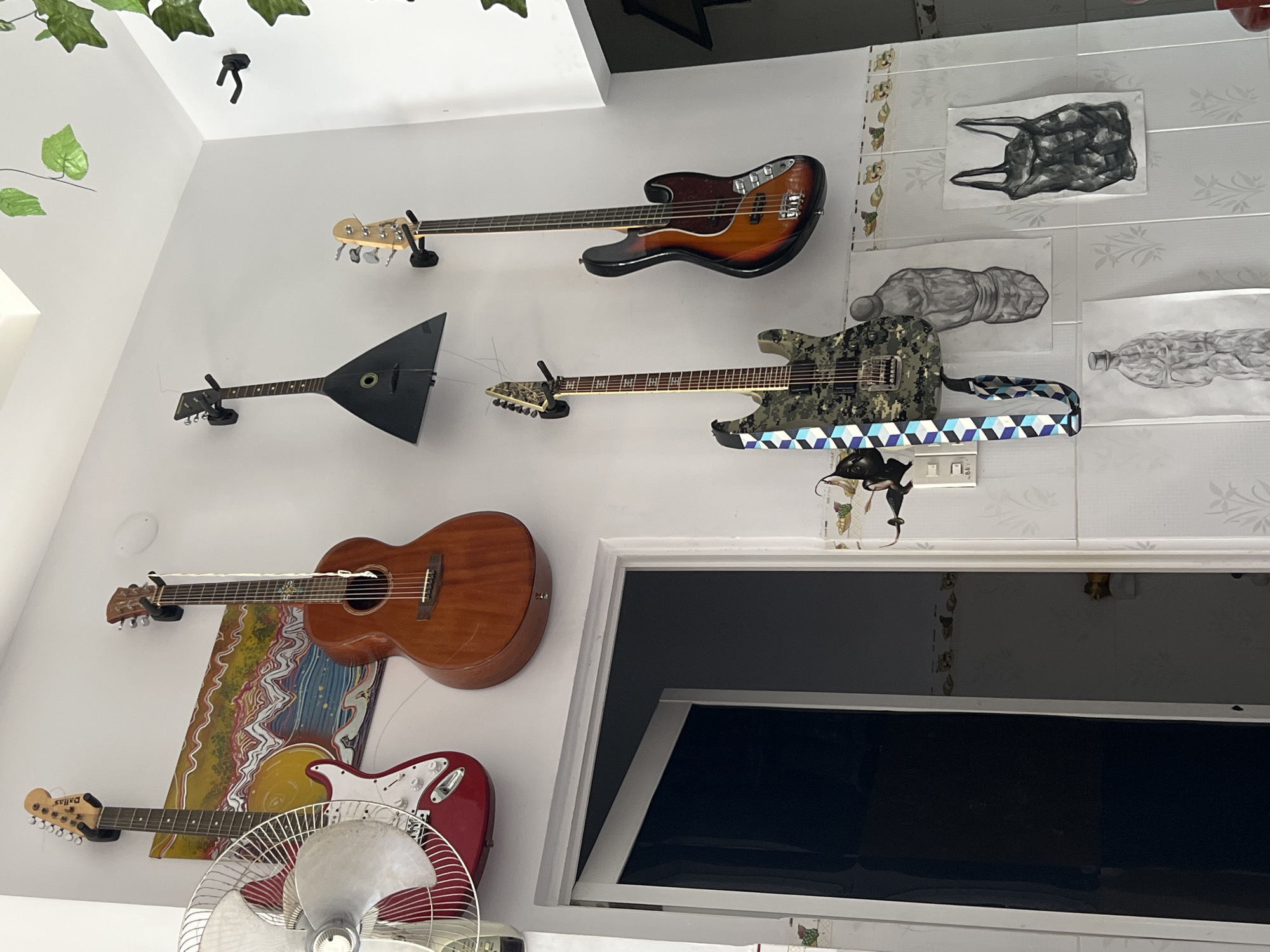 This image displays a brightly lit indoor wall section, identified as being within a residence in Nha Trang, Vietnam. The wall is predominantly white, featuring a lower wainscoting composed of white ceramic tiles adorned with a repeating pattern of green leaves and brown mushroom-like motifs.
Five stringed musical instruments are mounted on the white wall using black wall hangers. From left to right: a red electric guitar with a white pickguard and "Dallas" branding on the headstock, a brown acoustic guitar, a black triangular-bodied string instrument (resembling a Balalaika), a camouflage-patterned electric guitar equipped with a blue and black geometric strap, and a four-string bass guitar with a sunburst finish. A multi-colored abstract painting, featuring wavy patterns in yellow, orange, blue, and white, is horizontally mounted behind the red electric and brown acoustic guitars.
A white electric desk fan with a metal cage and red body is mounted on the wall in the upper left, partially obstructing the red electric guitar. Below the camouflage electric guitar and the bass, four monochrome drawings or prints are affixed to the tiled wall: two depict crumpled, textured objects resembling bottles or figures, and two depict crumpled black plastic bags. A small black feline-like silhouette ornament is attached to the wall below the camouflage guitar's headstock. White electrical wall switches are situated below this ornament. Artificial green ivy-like foliage is draped from the white ceiling in the upper right. An open doorway on the left reveals a dark interior space. No people are present in the scene, and no actions or interactions are depicted.