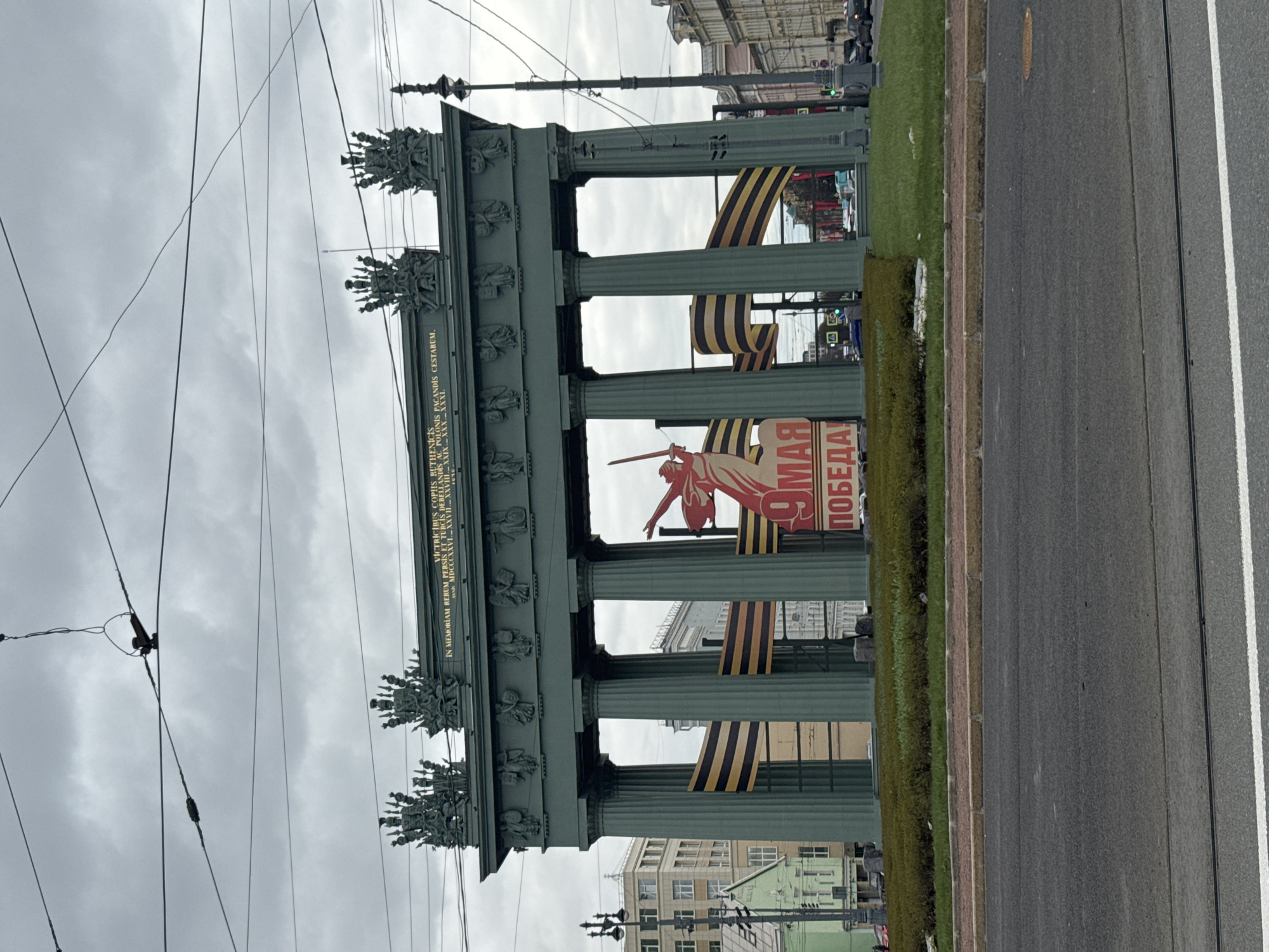 An outdoor, eye-level shot captures a large, ornate triumphal arch or colonnade under an overcast sky in Saint Petersburg, Russia. The multi-columned structure, likely the Narva Triumphal Arch, is made of dark stone or metal and features intricate sculptural decorations along its frieze and top.
A long Latin inscription is visible on the entablature, partially reading: "VICTRICIBUS COPIIS RUTHENICIS POLONIS PACANDIS CESTARUM. IN MEMORIAM RERUM PERSIS ET TURCIS DEBEVANDIS AL POLONIS PAGANDIS ANNIS".
Between the columns and in front of the arch, large red banners are displayed, featuring a stylized red figure holding a sword and the prominent Russian text "9 МАЯ ПОБЕДА!" (9 May Victory!). Numerous black and orange striped St. George ribbons, symbols of military valor and commemoration, are also draped and attached to the columns.
The arch stands alongside a multi-lane asphalt road with a grassy median strip. Overhead power lines crisscross the cloudy sky above. Buildings are visible in the background, confirming an urban setting. The scene depicts a public space decorated for the celebration of Victory Day.