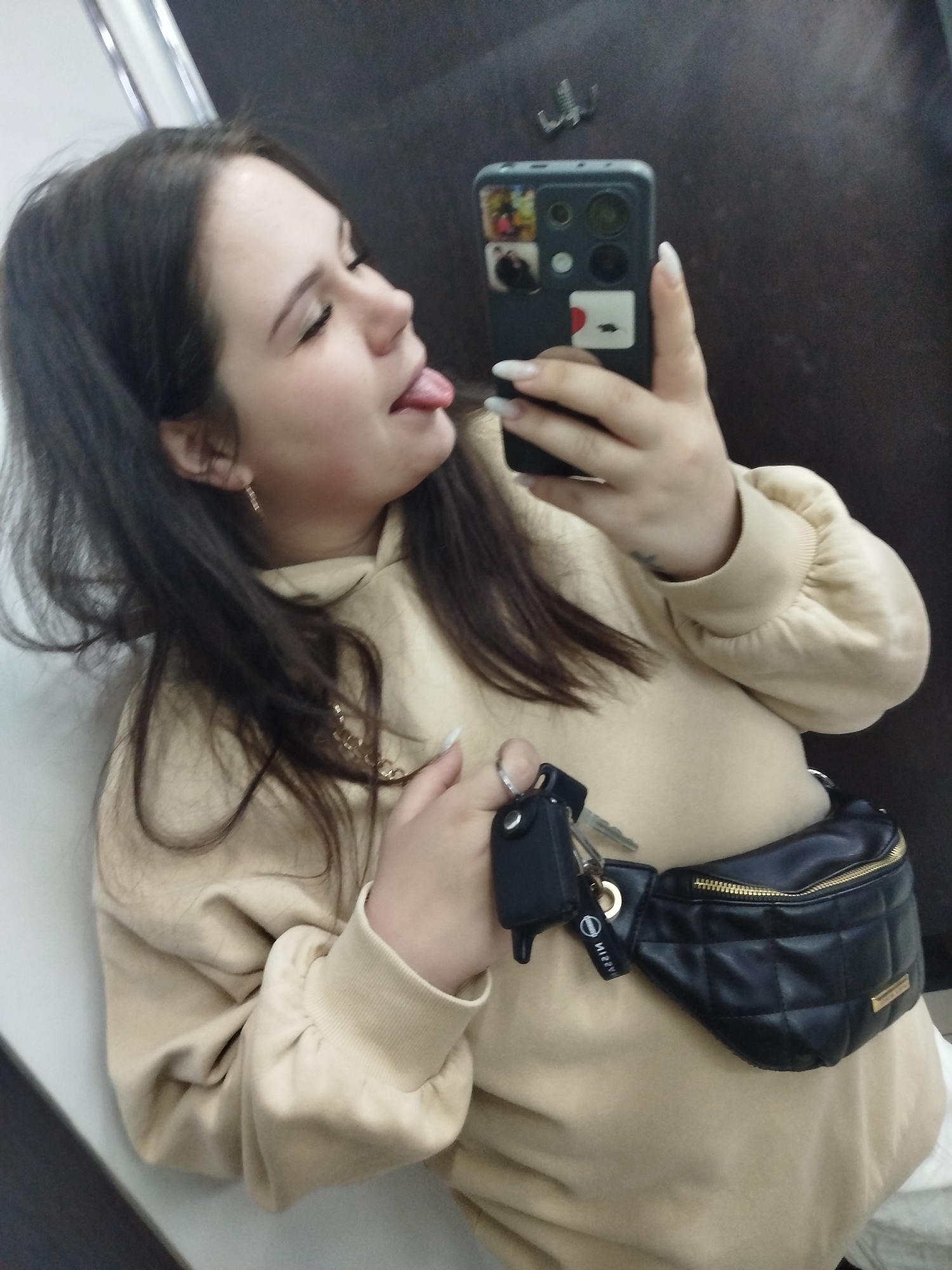 A woman is taking a selfie with a black smartphone with multiple stickers on the back. She is sticking her tongue out. She has long dark hair, fair skin, and is wearing a beige sweatshirt. A black quilted fanny pack with a gold zipper is around her waist and keys are hanging from it. The location appears to be inside a room with a dark brown wall and a white doorframe. The room could be located in Chaikovsky, Russia.