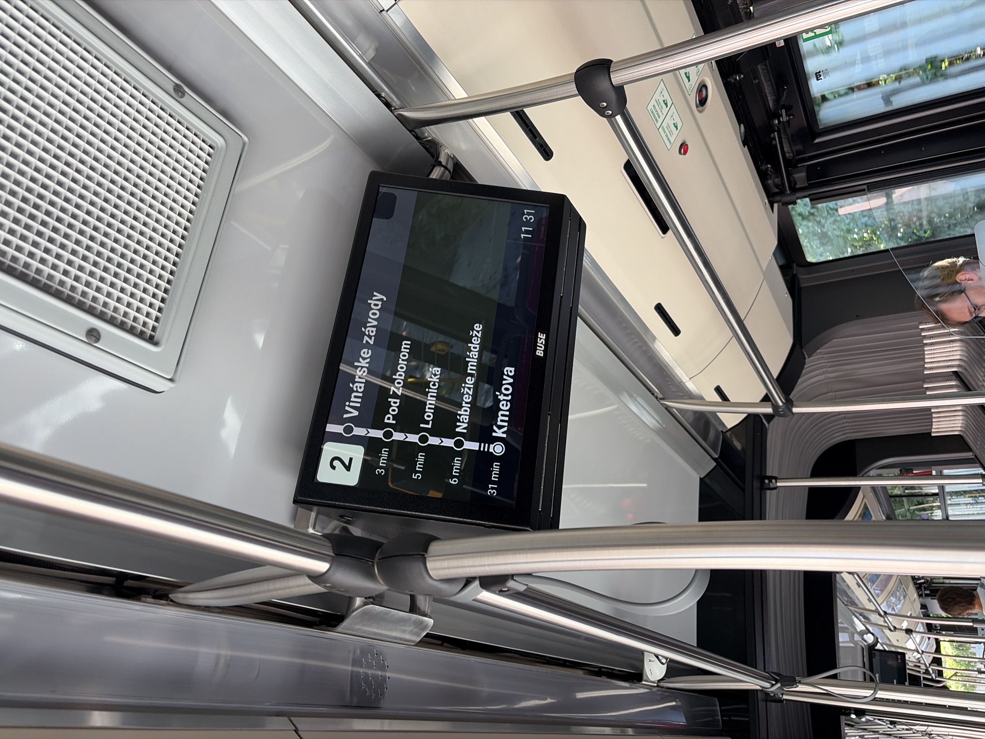 The image shows the interior of a public transport vehicle, likely a bus or tram, with a digital display screen mounted overhead. The screen shows route information, with the route number "2" prominently displayed. Below the route number, a list of stops and estimated arrival times is visible. The stops include "Vinárské závody", "Pod Zoborom", "Lomnická", "Nábrežie mladéže", and "Kmetová". The times listed are 3 min, 5 min, 6 min, and 31 min respectively. The screen also displays "11 31" which could be the current time. The interior of the vehicle features metal support poles and overhead compartments. Through the windows, greenery is visible, suggesting an outdoor environment. The overall scene depicts a typical urban public transportation setting in Nitra, Slovakia, likely on a route that serves the mentioned locations, which are all within or in the vicinity of Nitra. There are no identifiable people clearly depicted in the foreground.