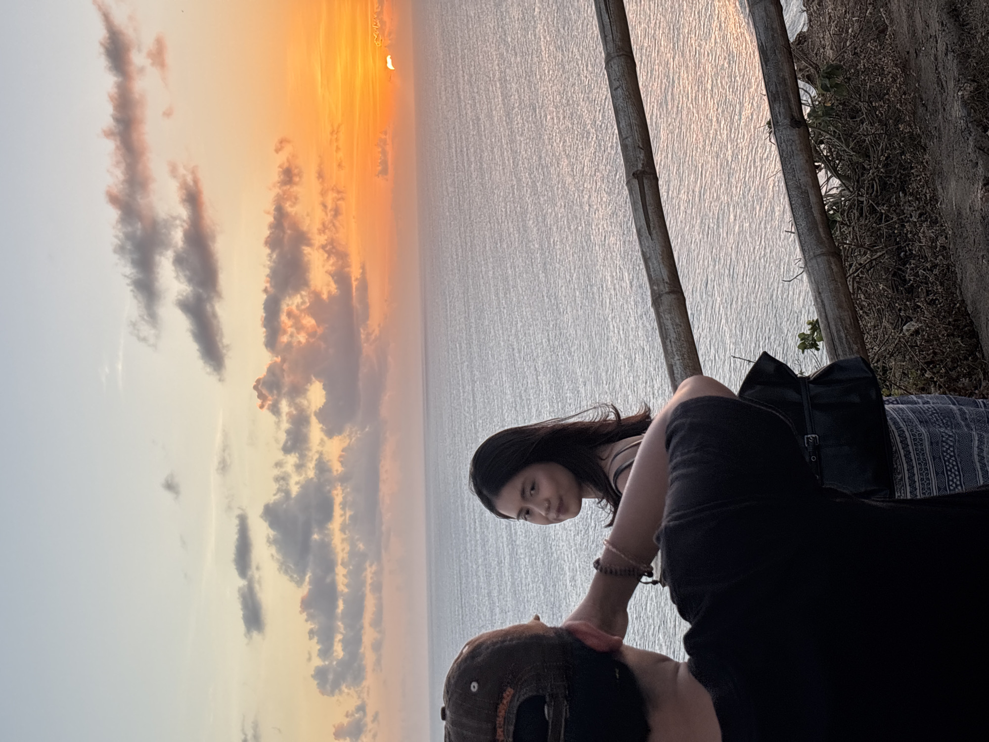 Two individuals, one facing away from the camera and another facing towards it, are positioned on a cliff overlooking the ocean at sunset. The person in the foreground, seen from the back, wears a black t-shirt and a baseball cap. The second individual, a young woman with dark hair, is looking over her shoulder towards the camera, a slight smile on her face. She is wearing a beige spaghetti strap top. A dark handbag is slung over her shoulder. A wooden railing made of bamboo runs across the middle ground of the image, separating the elevated viewpoint from the sea. The ocean stretches out to the horizon, reflecting the warm hues of the sunset. The sky is painted with a gradient of soft blues, oranges, and pinks, with scattered clouds adding texture. The overall scene suggests a serene moment of enjoying the coastal sunset in Kuta Selatan, Indonesia, likely a popular scenic spot.