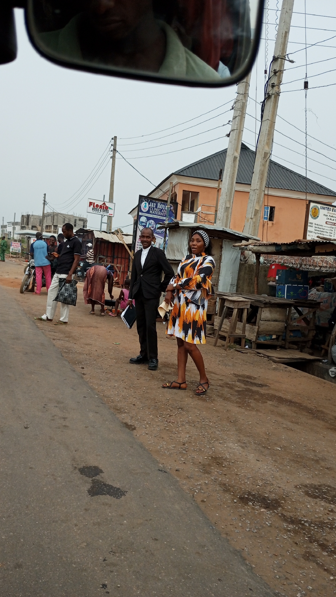 The image shows a street scene in Adankolo, Nigeria. Two individuals, a man in a black suit holding a black folder and a woman in a colorful dress and head covering, are standing on a dirt patch next to a paved road. Behind them, there is a collection of makeshift stalls and buildings, suggesting a market or commercial area. Several people are standing and interacting near the stalls. Signage for "FLORIC HOTELS & SUITES" and "JAY KOYA CLINIC ANI MATERNIT" is visible in the background. Utility poles and wires are overhead. The weather appears overcast. Part of a car mirror is visible in the upper left corner of the frame.