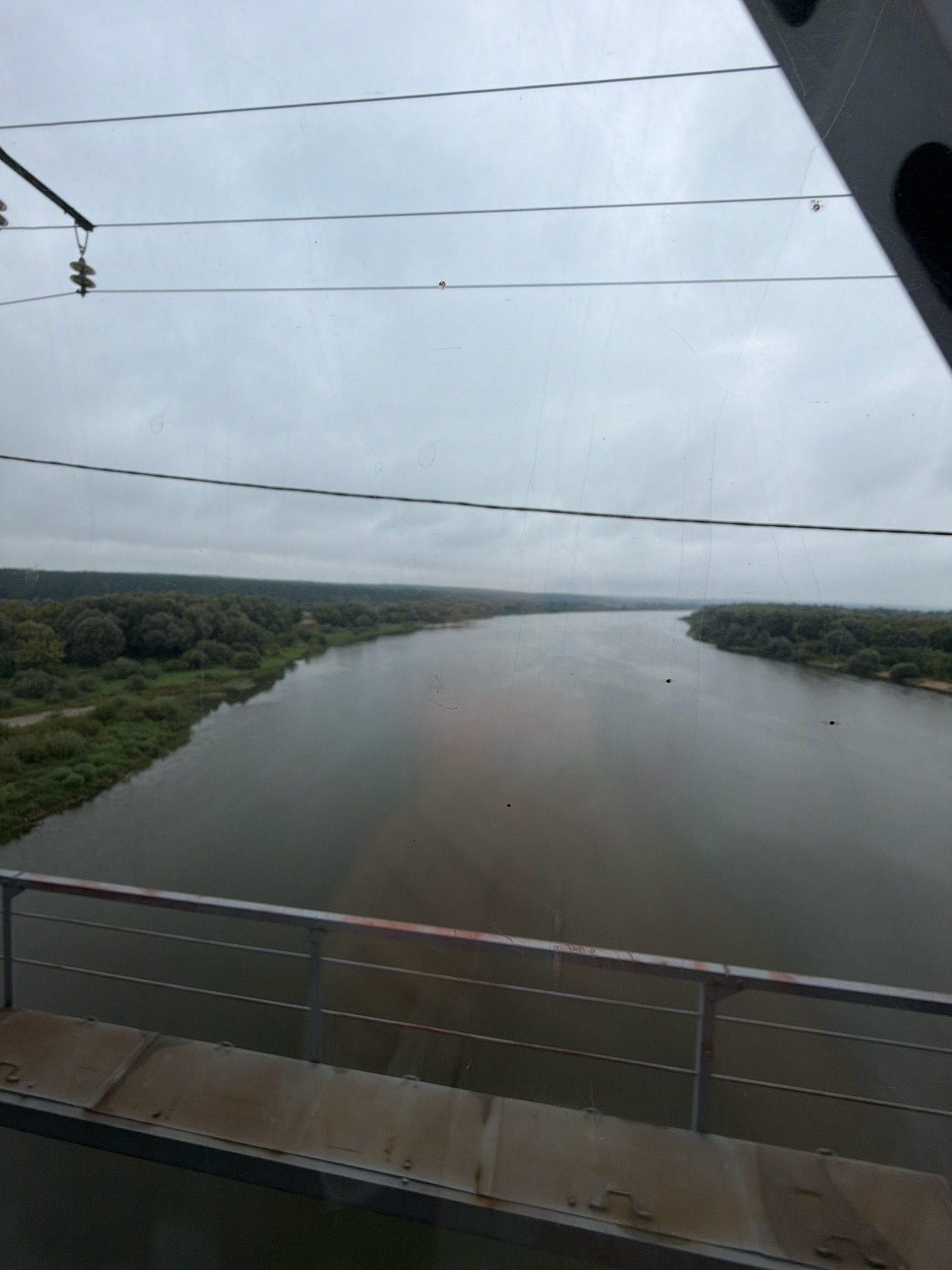 The image depicts a wide, calm river with tree-lined banks under an overcast sky, viewed from a bridge. The bridge railing is visible in the foreground, with a metal walkway. Electrical wires and insulators are strung across the sky. The scene is consistent with the Oka River in Kashira, Russia, known for its bridges and scenic river views.