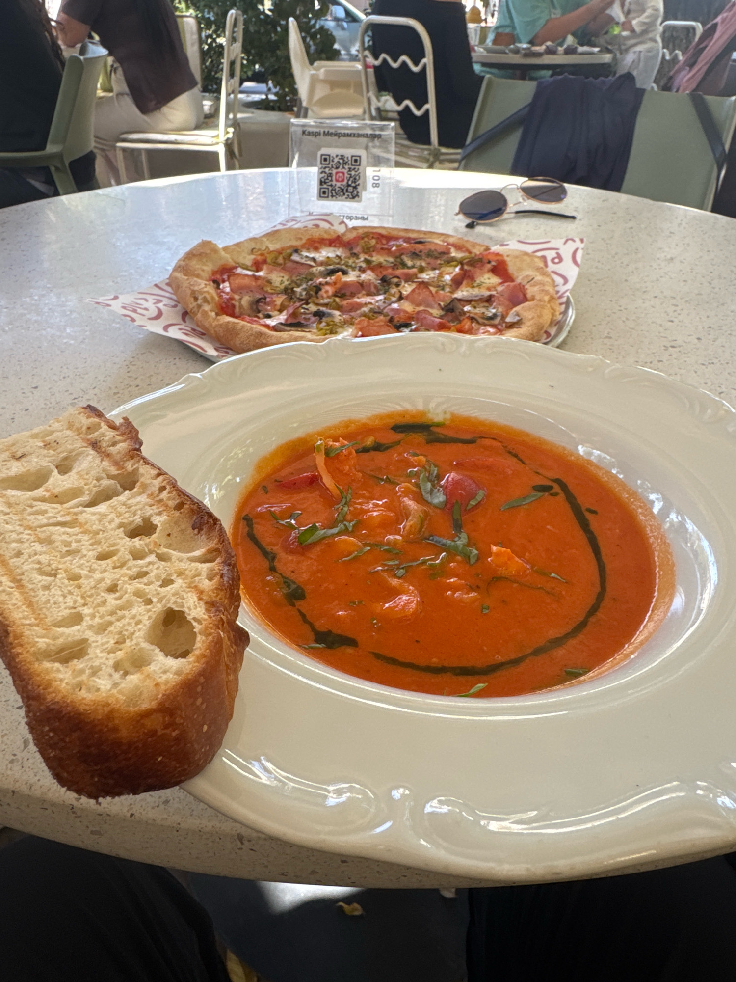 A close-up, eye-level shot captures a meal served at an outdoor dining establishment, likely in Almaty, Kazakhstan, given the presence of a QR code for "Kaspi.kz" which is a prominent Kazakhstani financial technology company. In the foreground, a white bowl filled with a vibrant red soup, possibly tomato soup, is garnished with fresh green herbs and a swirl of dark oil. To the left of the soup, a thick slice of crusty bread is positioned, its airy interior and golden-brown crust visible.
Behind this dish, a thin-crust pizza rests on a decorative paper liner. The pizza appears to be topped with mushrooms, ham, and possibly peppers. In the middle ground, on a speckled tabletop, a pair of sunglasses with dark lenses and a thin gold frame lies beside the pizza. Further in the background, out of focus, several people are seated at tables, with some appearing to be diners and others staff members. Chairs and other tables are visible, suggesting a restaurant or cafe setting. The lighting indicates it is daytime, with sunlight illuminating the scene.