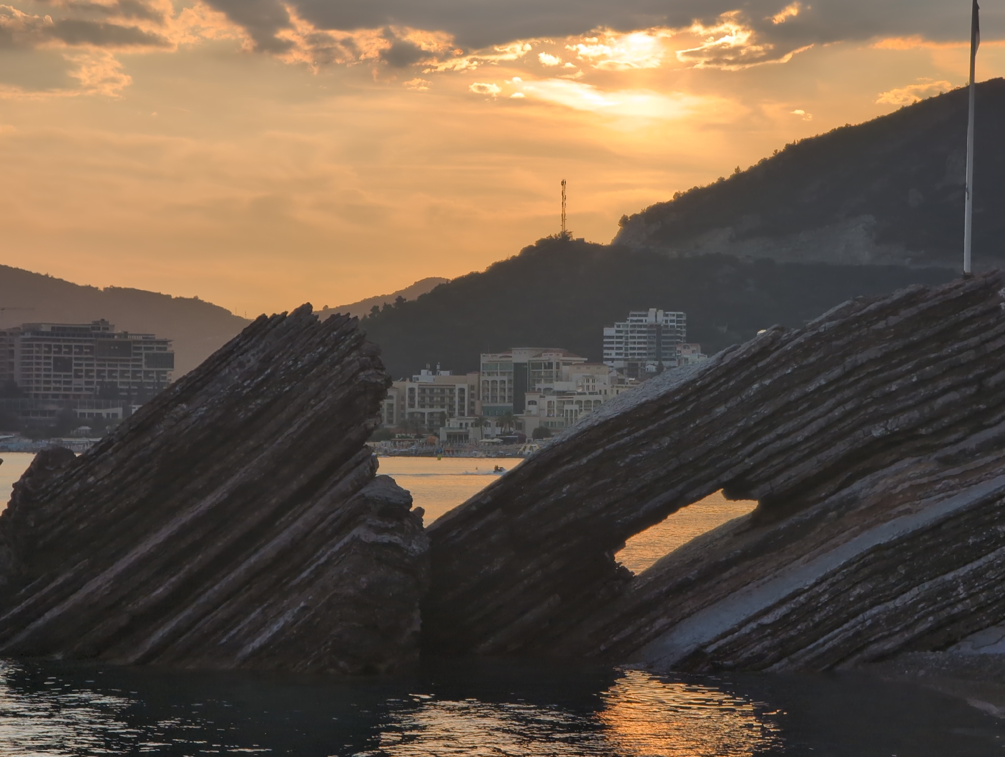 This media file depicts a scenic coastal view at sunset in Rafailovići, Montenegro. The foreground is dominated by rugged, layered rock formations jutting out of the water. The rocks are dark gray with distinct horizontal striations, and one section has a natural archway. The water surrounding the rocks reflects the warm, golden hues of the sunset, creating shimmering patterns.
In the midground, a town is visible nestled along the coastline, with numerous buildings, including hotels and apartments, featuring white facades and visible balconies. Further back, the dark silhouettes of hills and mountains rise, adding depth to the landscape. A tall, slender tower, possibly a telecommunications mast, is visible on one of the hills. A flagpole stands prominently on the right side of the frame.
The sky is filled with dramatic clouds illuminated by the setting sun, which appears as a bright, diffused glow behind a band of clouds. The overall atmosphere is serene and tranquil, characteristic of a beautiful sunset. A jet ski can be seen in the water near the town, indicating some recreational activity. There is no visible text in the image. The time of day is clearly sunset, with the warm, soft light casting long shadows and bathing the scene in golden and orange tones. The weather appears clear, with the clouds adding visual interest to the sky. The presence of the town and the hilly terrain strongly suggest a coastal location.