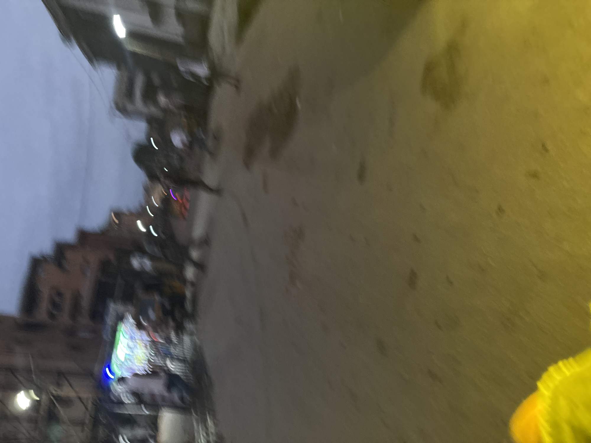 The image depicts a street scene in Onitsha, Nigeria, under a twilight sky. The scene is blurred due to motion, creating streaks of light from what appear to be streetlights and illuminated signs. Several figures are visible, walking or standing on the street, some appearing to be interacting or in motion. Buildings line the street, with some structures showing signs of illuminated windows or storefronts. The ground is a paved surface, possibly asphalt, with some darker patches that could indicate wet areas or shadows. In the foreground, a blurred section of bright yellow material is visible, possibly clothing or an object. The overall impression is of a busy, active street at dusk.
