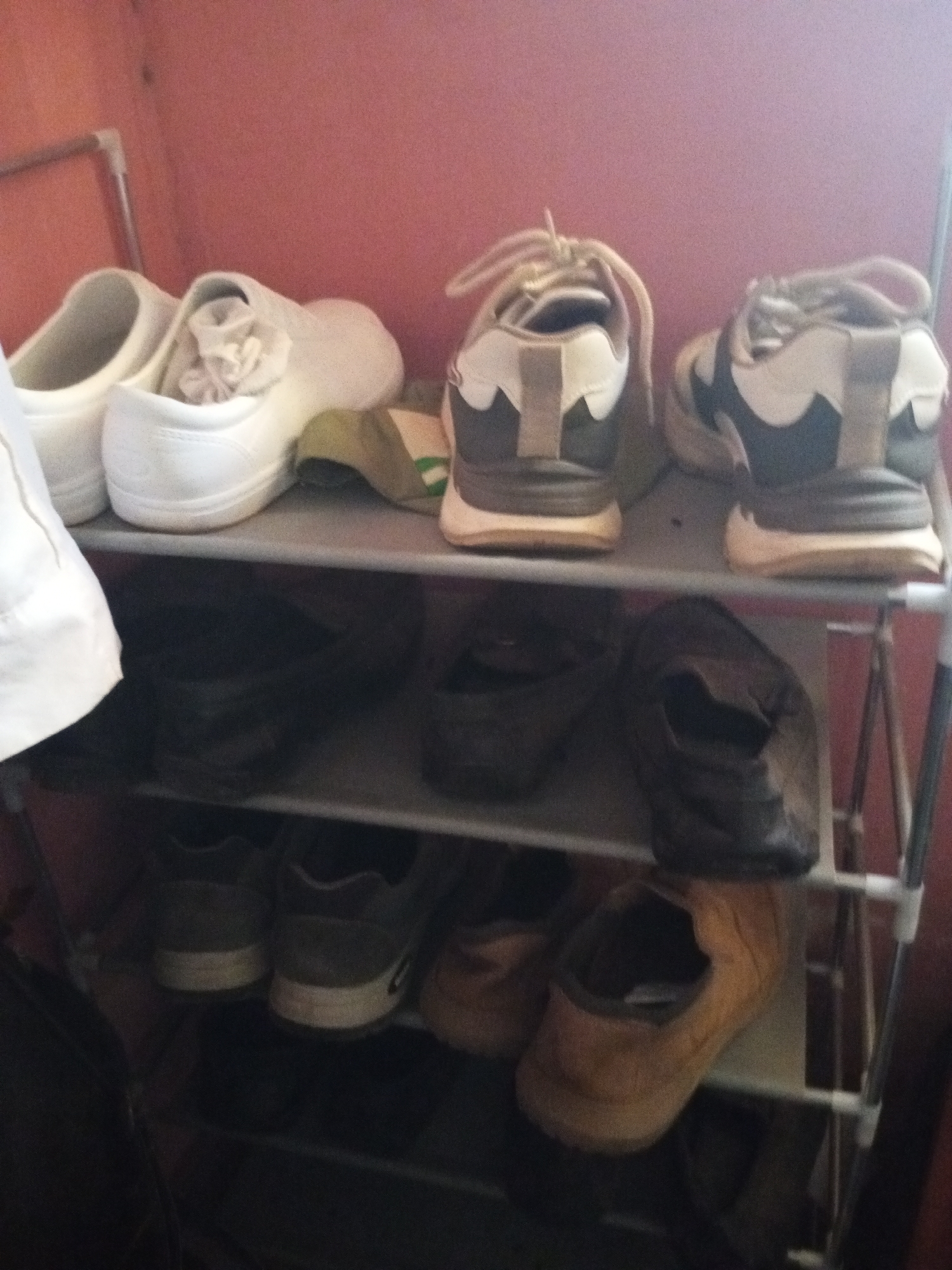 The image shows a four-tiered shoe rack filled with various footwear. The shoe rack is positioned against a pinkish wall. On the top shelf, there are two white slip-on shoes, one with a pinkish fabric insert, and a pair of grey and white athletic shoes. The second shelf contains two pairs of dark-colored shoes, possibly sneakers or boots. The third shelf has a pair of grey athletic shoes and a pair of tan slip-on shoes. The bottom shelf is mostly obscured by shadows but appears to contain dark-colored footwear. The overall lighting in the image is dim. The scene is not discernable as being in Jalingo, Nigeria.