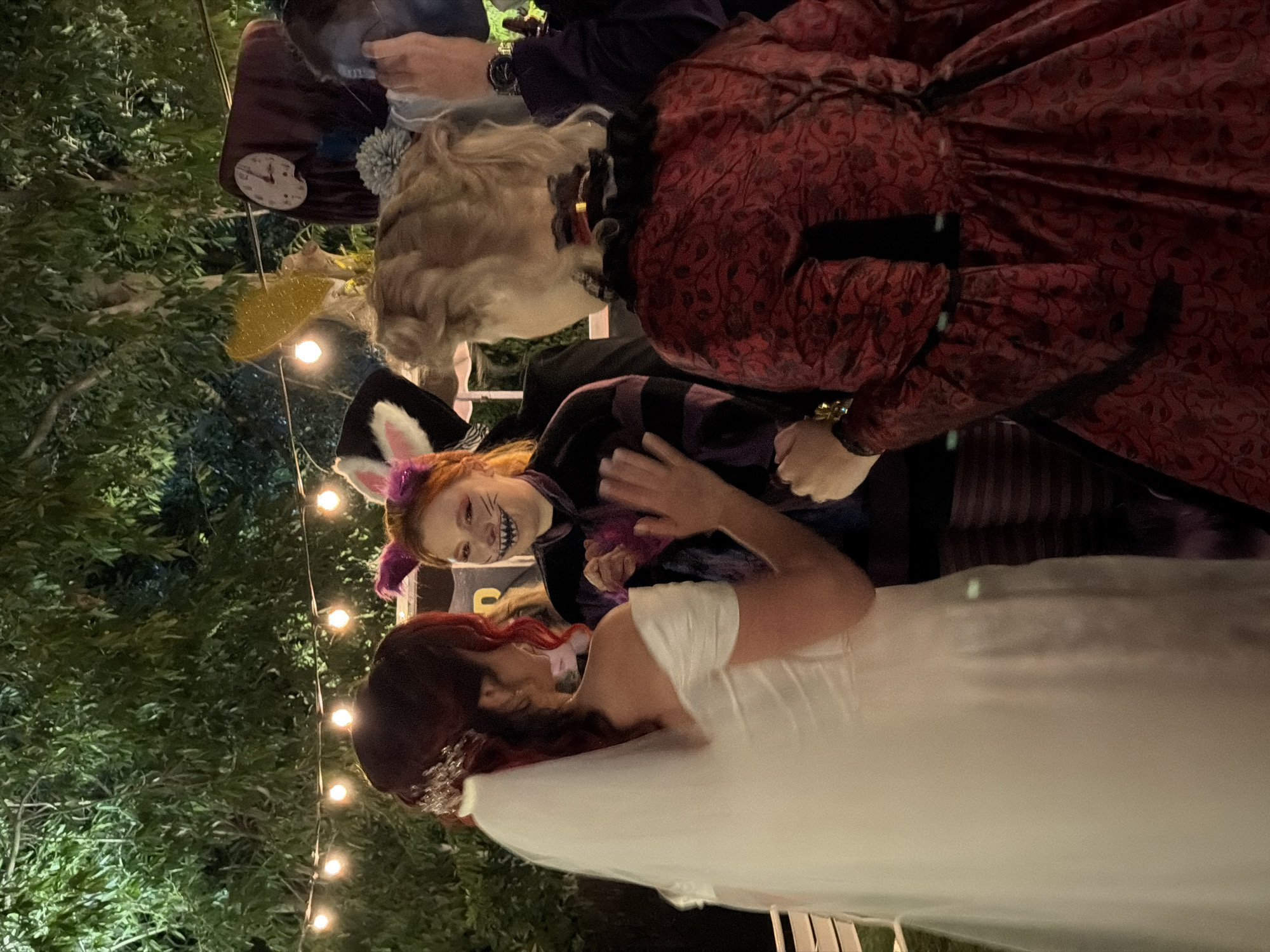 This evening photograph, taken in Nir Eliyahu, Israel, depicts a lively outdoor event, likely a themed party or wedding reception, illuminated by warm string lights. The background consists of dark green foliage.
In the center of the frame, a person dressed as the Cheshire Cat from Alice in Wonderland is prominently featured. They wear a purple and black striped top, purple cat ears, and have their face painted with whiskers and a wide, mischievous grin. They are looking towards a person in the foreground who has vibrant red hair and is wearing a white dress, possibly a bridal gown, along with a sparkling hair accessory.
Behind the Cheshire Cat, another individual is visible in a dark red dress with an ornate, possibly Victorian-era, pattern. Further back and to the left, parts of other costumed individuals can be seen; one appears to be wearing a light-colored wig and holding a small clock face, suggesting the character of the White Rabbit. The overall mood is festive and whimsical, characterized by the elaborate costumes and the soft glow of the artificial lighting. No text is visible in the image.