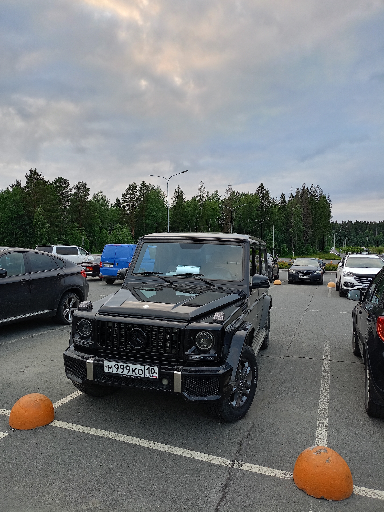 A black Mercedes-Benz G-Class SUV is parked in a paved lot with a wooded area in the background under a cloudy sky. The SUV is facing forward, with its front grille and headlights clearly visible. The license plate reads "M 999 KO 10" and includes "RUS" for Russia. Several other vehicles, including a black BMW X6, a blue van, a white SUV, and a black sedan, are parked in the vicinity. Orange spherical concrete barriers are situated in front of the parking spaces. The scene appears to be a public parking area.