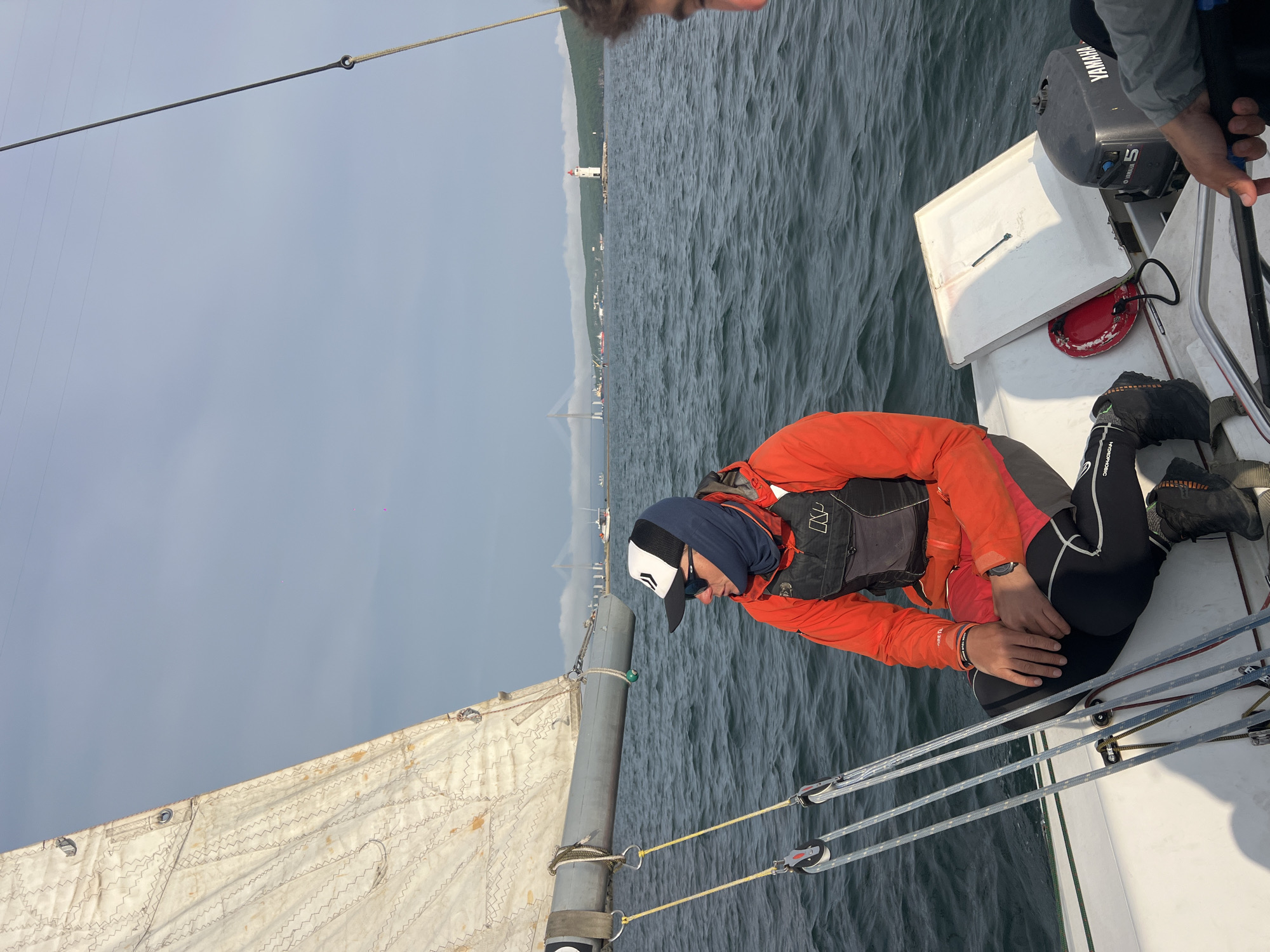 On a sailboat in Vladivostok, Russia, a man wearing an orange jacket and a baseball cap is kneeling. The sailboat is equipped with a Yamaha engine. The water is choppy and the sky is overcast. In the background, there is a lighthouse and a bridge.