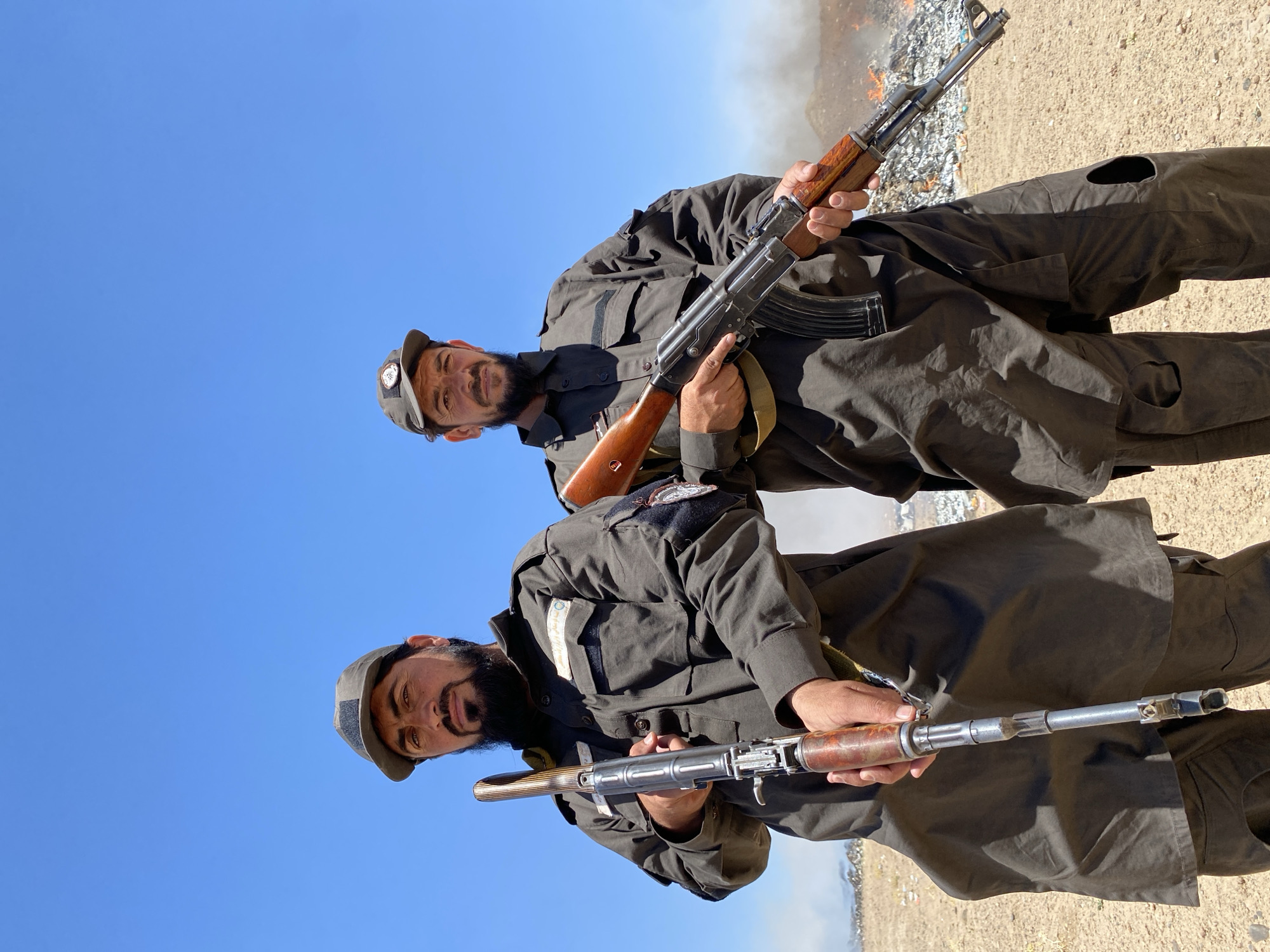 Two men, each wearing dark military-style uniforms and caps, stand holding AK-47 rifles in what appears to be a desert environment. The man on the left, with a beard and a darker cap, holds his rifle vertically in front of him. The man on the right, also bearded and with a lighter cap, holds his rifle horizontally across his body. Behind them, a large pile of burning trash or debris is visible, with smoke rising into the clear blue sky. The ground is sandy and dry. The image is set in Mandisar, Afghanistan.