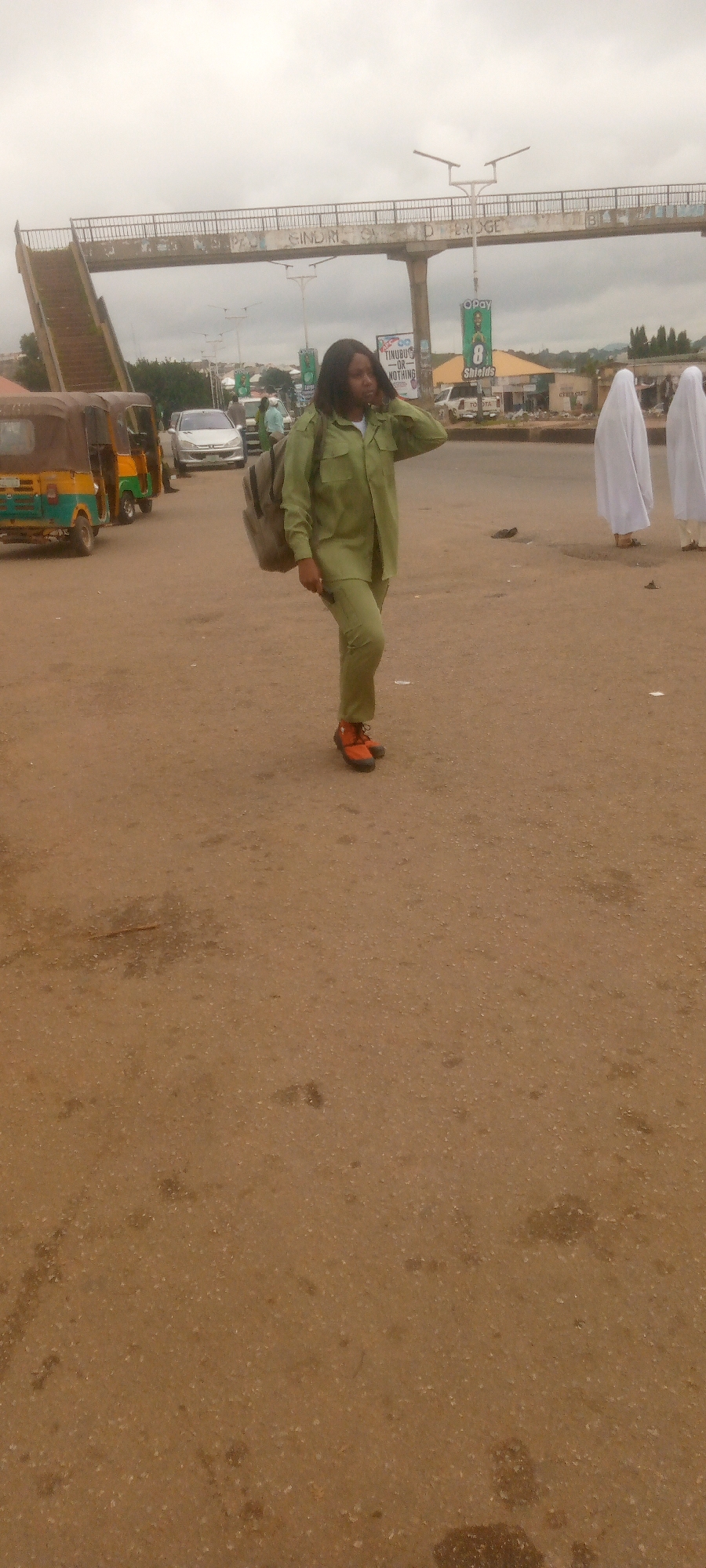 A young woman, dressed in a light green uniform and carrying a backpack, stands on a dusty road in an urban area. She has her right hand raised to her ear, appearing to be on a phone call. Her orange shoes are visible. In the background, a pedestrian overpass with graffiti stretches across the sky. Several vehicles, including a yellow and green motorized tricycle, a silver car, and other cars, are present on the road. Two individuals in white coverings are visible further in the background. Several billboards and signs are also visible in the distance, including one with the OPay logo and another that reads "TINUBU - OR - NOTHING". The sky is overcast.