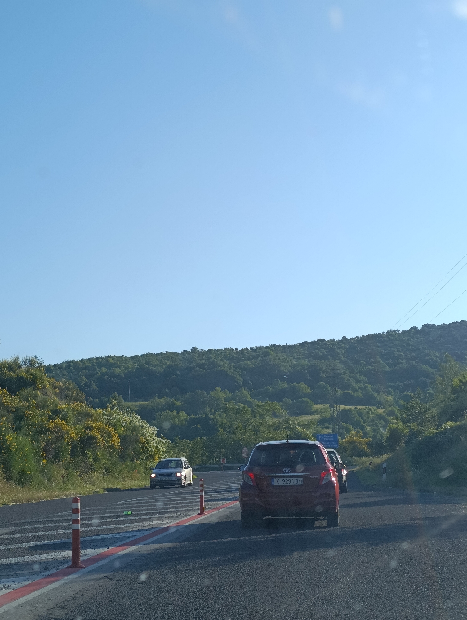The image captures a view from a vehicle traveling on a road near Cherna Skala, Bulgaria. The road is paved and has multiple lanes, with white and red reflective markers delineating the lanes. In the foreground, a red Toyota Yaris is visible from behind, with a Bulgarian license plate "K 9291 BH". To the left of this vehicle, another car, possibly a silver minivan, is seen from the front, with its headlights on. The road curves to the right. The environment consists of a steep, tree-covered hillside to the right, with dense green foliage and some yellow flowering bushes on the left side of the road. The sky is clear and blue, indicating a sunny day. A blue road sign with white lettering indicating directions to "Varna", "Silistra", and "Veliko Tarnovo" is visible in the distance. The overall scene depicts a scenic drive on a highway or main road in a rural, hilly region of Bulgaria.