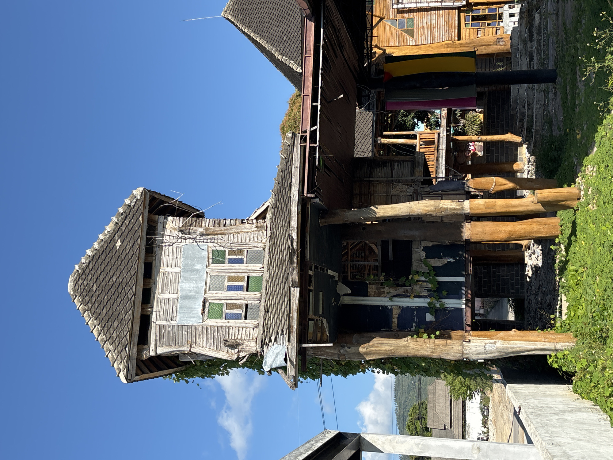 Baan Chaweng, Thailand: A multi-level wooden structure is supported by tree trunk columns. The structure appears aged and partially dilapidated, with exposed wood and sections missing or damaged. Some windows have multi-colored panes. The structure is surrounded by foliage. The sky is clear blue with some clouds visible in the background.