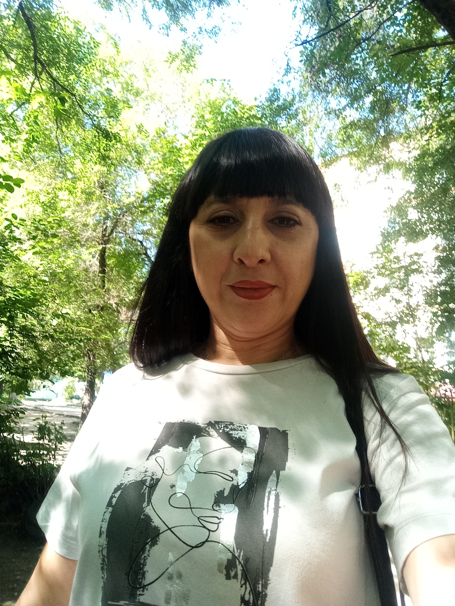 A woman with dark, straight hair and bangs takes a selfie outdoors. She is wearing a white t-shirt with a black and gray abstract design. She has on red lipstick. The background is filled with lush green trees and dappled sunlight, suggesting a park or a wooded area in Belogorsk, Russia. The lighting indicates it is daytime. The woman is looking directly at the camera.