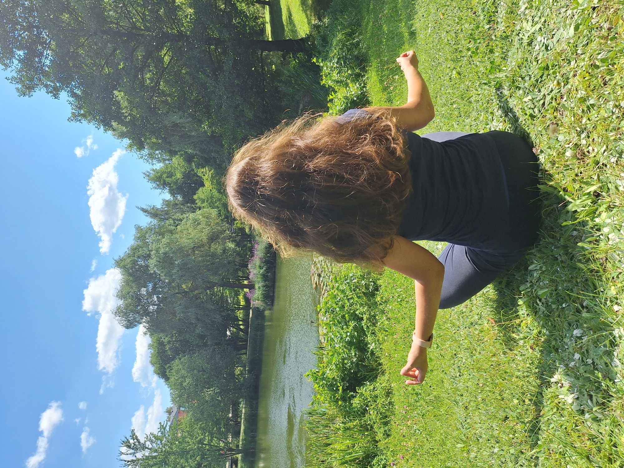 A woman with long, wavy brown hair is meditating outdoors on a grassy hill. She is sitting cross-legged, facing away from the camera, with her hands resting on her knees in a meditative pose. Her eyes are closed, and she appears to be relaxed. The background features a body of water, possibly a lake or pond, with lush green trees and vegetation surrounding it. The sky is blue with a few white clouds. The overall scene suggests a peaceful and serene environment. This image was taken in the city of Wolkenstein, Germany.