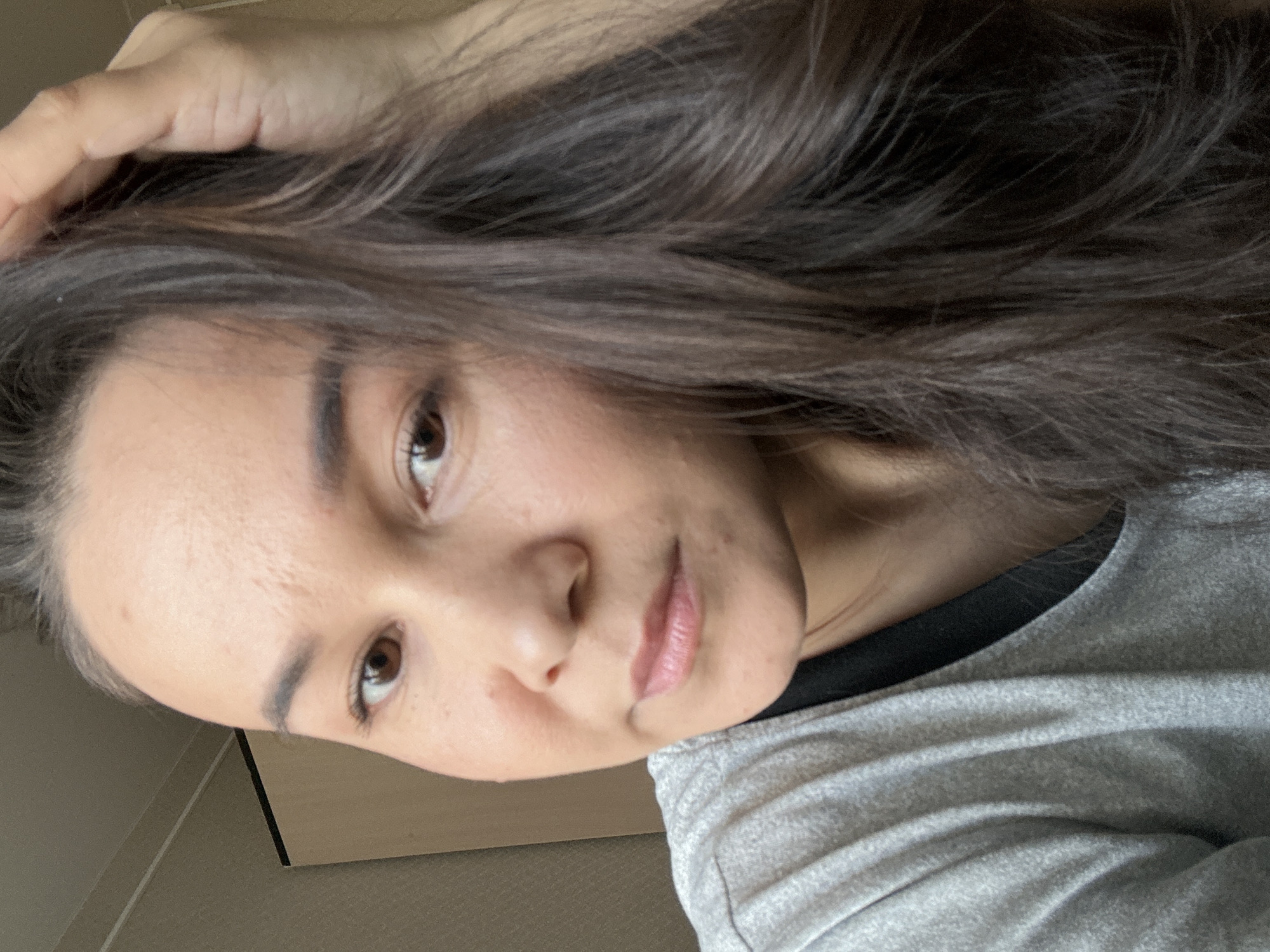 A medium, close-up shot of a woman with dark hair, brown eyes, and fair skin. She is wearing a gray shirt with a black collar. Her left hand is touching her hair. The background shows a cream-colored door or wall and it is impossible to tell if this is taken in the city of Uray, Russia.