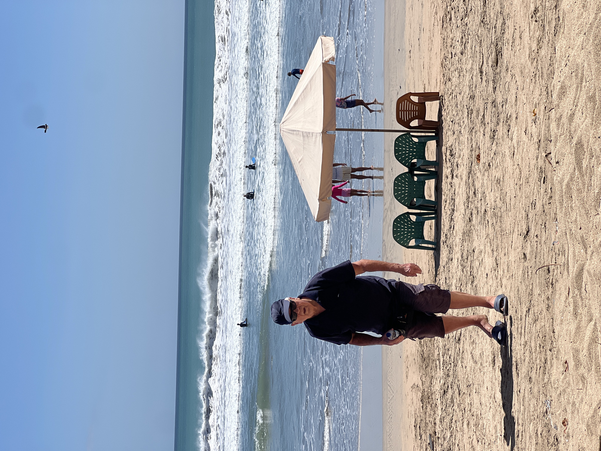 In Kuta, Indonesia, an elderly Caucasian man wearing a dark blue polo shirt, grey cargo shorts, a dark blue baseball cap, and sunglasses walks on a sandy beach towards the camera. He holds a clear plastic bottle of water in his right hand. Behind him, a beige beach umbrella shades four plastic chairs, three green and one brown. In the background, the turquoise ocean meets a bright blue sky, with white waves crashing ashore and several surfers in the water. A bird flies high in the sky.