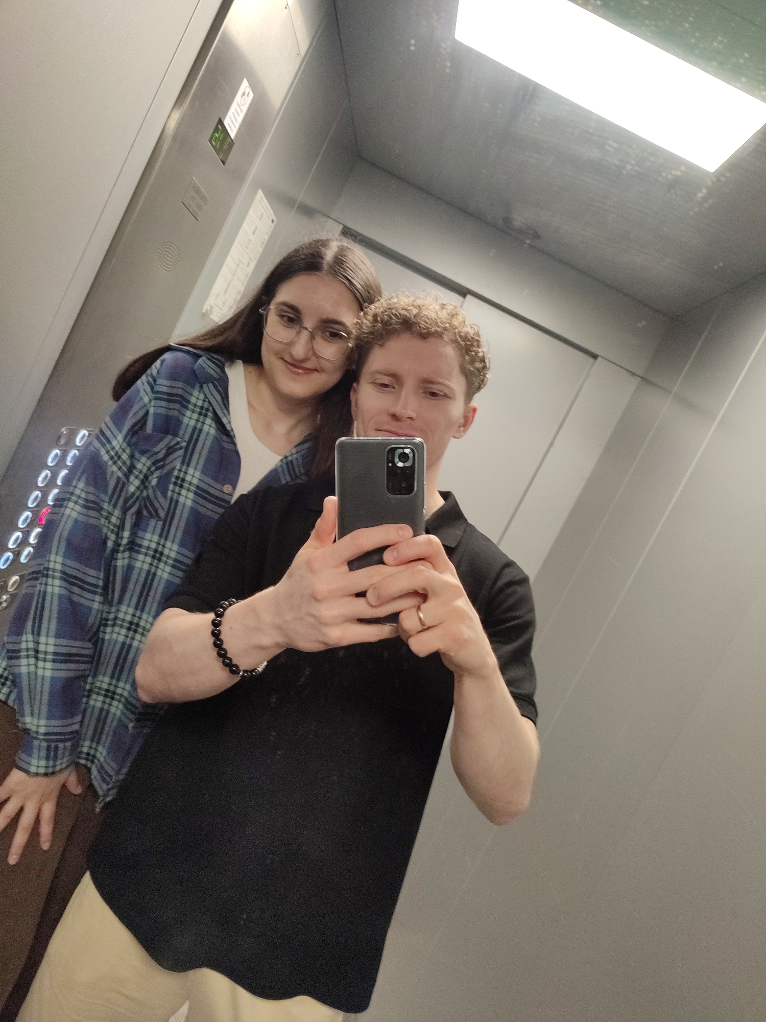 A young Caucasian couple is taking a mirror selfie in an elevator. The man, in the foreground, is wearing a black polo shirt and light-colored pants. He has curly blonde hair and is wearing a ring on his left ring finger. He is holding a smartphone to take the picture. The woman, behind him, has long dark hair and is wearing glasses and a blue and white plaid shirt over a white top. They are both smiling. The elevator has stainless steel walls and a bright overhead light. There are elevator buttons on the left wall.
