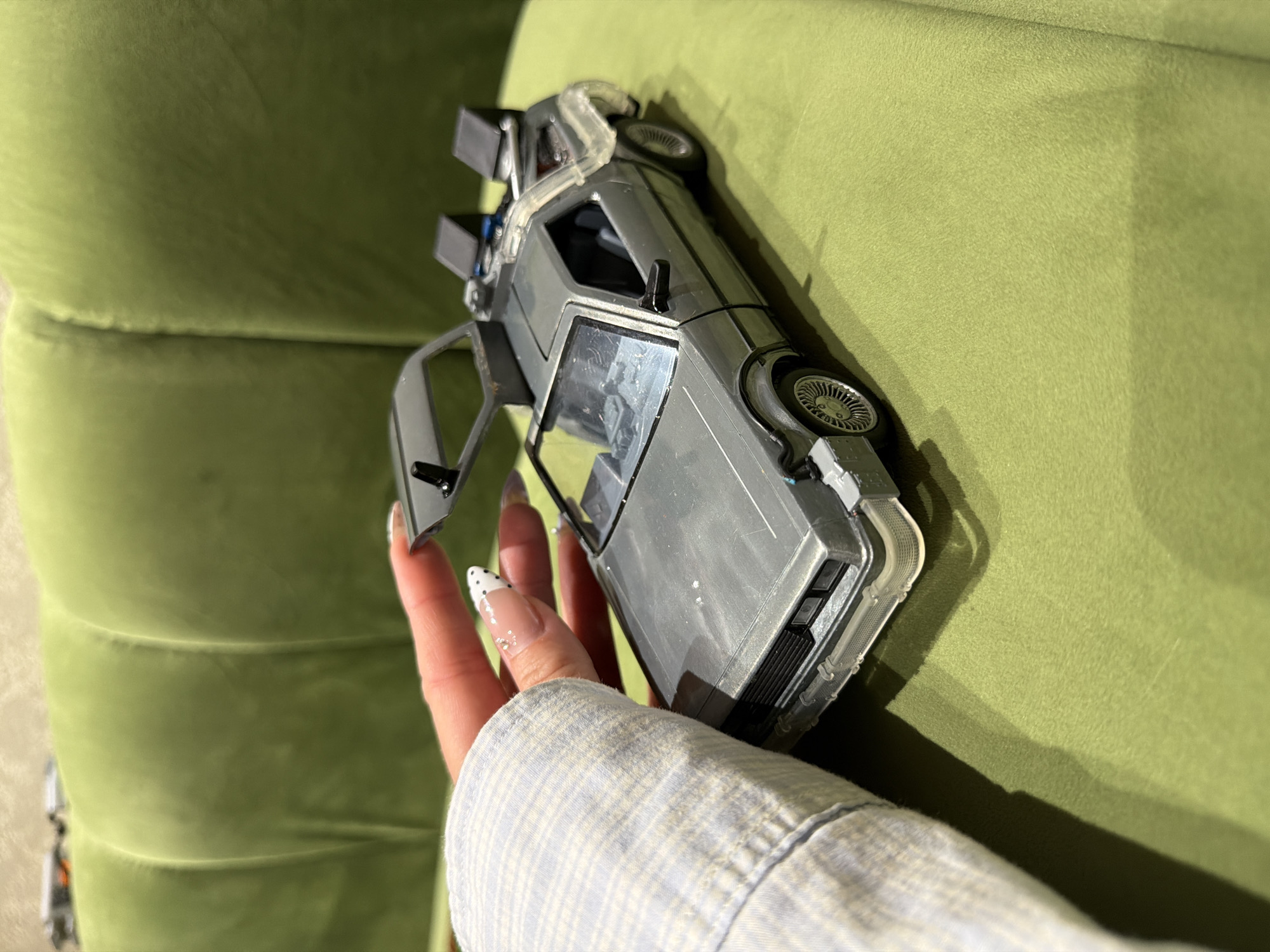 A person wearing a light blue and white plaid shirt is holding a silver toy DeLorean car with open gull-wing doors. The car is resting on a green, textured surface, likely a couch. The person's hand with long nails and a light manicure is visible. The location is not discernible from the image and therefore cannot be confirmed as Kuta Selatan, Indonesia.
