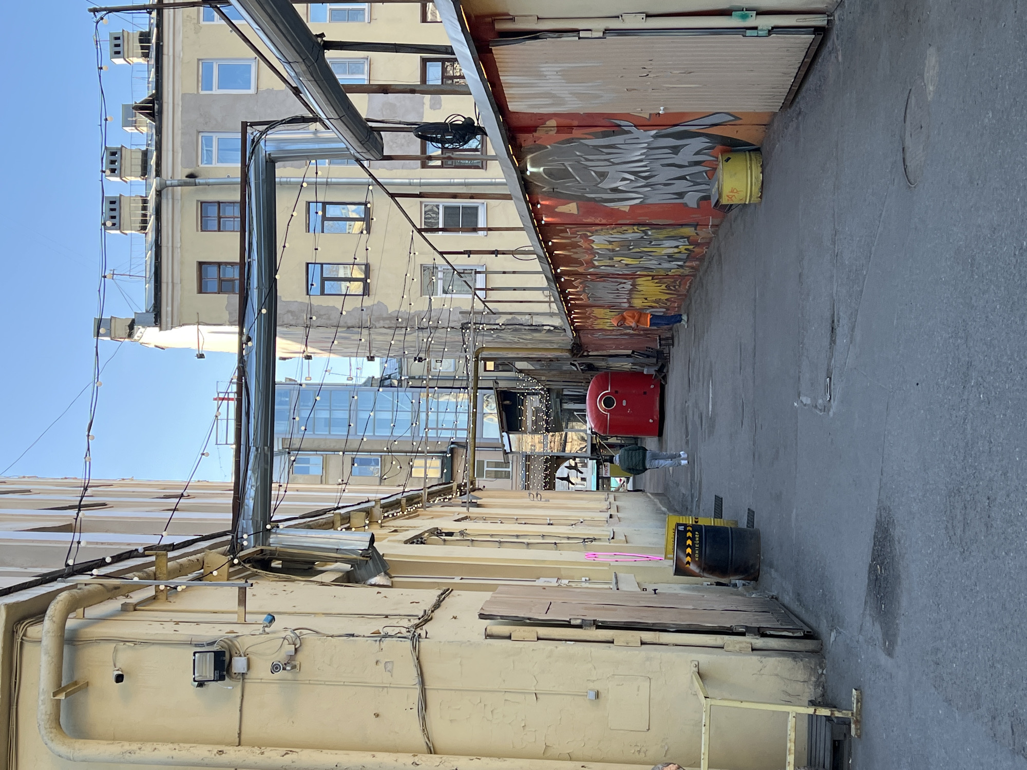 The image depicts an alleyway in Saint Petersburg, Russia. The alley is flanked by buildings on either side, one with a light yellow facade and the other constructed from shipping containers stacked on top of one another. The containers are decorated with graffiti. Overhead, strands of decorative string lights span the alley. A few pedestrians are visible in the distance. A red trash container is present on the street. Other objects are also visible, including a black barrel. The sky is clear and blue.