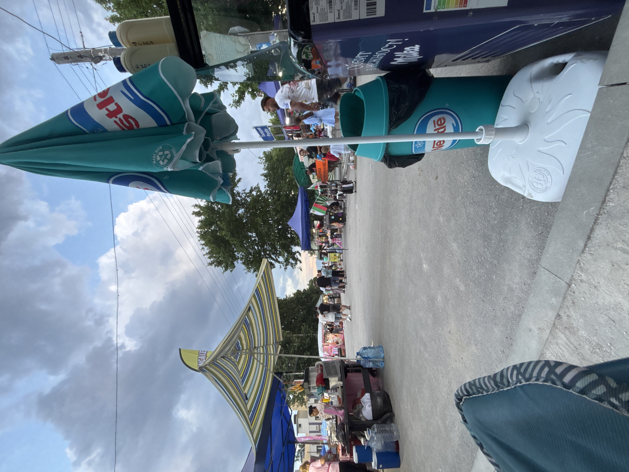 This image depicts an outdoor market or fair scene in Kostievo, Bulgaria, observed from a low vantage point, likely from a seated position. The setting appears to be an open-air pedestrian area with multiple vendor stalls under a partially cloudy sky. In the immediate foreground, a portion of a teal blue fabric chair with striped trim is visible on the bottom left. To the right, a large, closed teal blue sun umbrella with "Nestlé" branding in white and red lettering, along with a "MADE WITH RECYCLED PLASTIC" logo, is prominently displayed. The umbrella is secured by a white plastic base, resting on a light grey concrete or asphalt surface. Adjacent to the umbrella base is a teal green cylindrical bin also bearing the "Nestlé" logo, with a black trash bag visible at its opening. To the right of the bin, a dark blue commercial refrigerator or freezer is partially visible, displaying Cyrillic text "ОБИТ В НЕЖНОСТ! Milka" (Lived in tenderness! Milka) and an energy efficiency label. Two light-colored plastic bottles are placed on top of this refrigerator. In the midground to the left, a market stall features a distinctive striped canopy in shades of yellow, blue, green, and white. Above this canopy, a yellow banner-style flag is partially visible, showing the text "NCAKE" (possibly part of "PANCAKE"). Beneath the striped canopy, a woman in a light-colored shirt stands behind a counter, possibly engaged in food preparation or service, with various containers and a blender-like machine containing red liquid on the table. Several large plastic water bottles and blue coolers are arranged on the ground beneath the counter. A blue canopy from an adjacent stall is visible further to the left. The central and right midground shows a paved pathway extending into the distance, flanked by numerous market stalls. People are seen walking, standing, and interacting among the stalls. A man in a white t-shirt and dark shorts walks towards the right, followed by a young girl in a light blue dress. Near one of the stalls on the right, a signpost displays Cyrillic text "ПЛОВДИВ Plovdiv," indicating proximity or direction to the city of Plovdiv, and another sign below reads "БЪЛГАРСКА ЦАРЕВИЦА НА МАСА" (Bulgarian Corn on the Cob). A Bulgarian flag is visible at one of the stalls. Further down the pathway, a denser crowd of people is gathered around additional market setups. Trees line the background on both